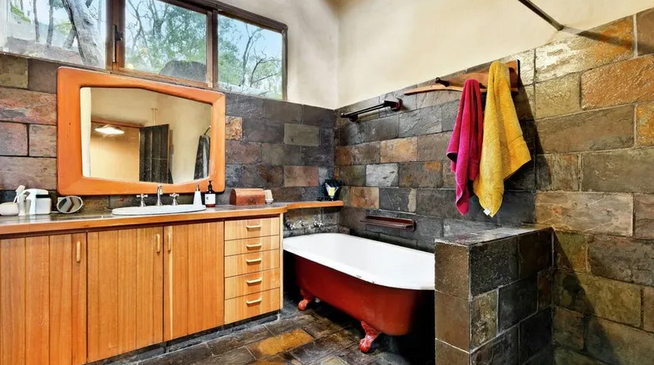 Bathroom with stone walls, a wooden vanity with a mirror, and a red clawfoot bathtub with towels hanging on a towel rack.
