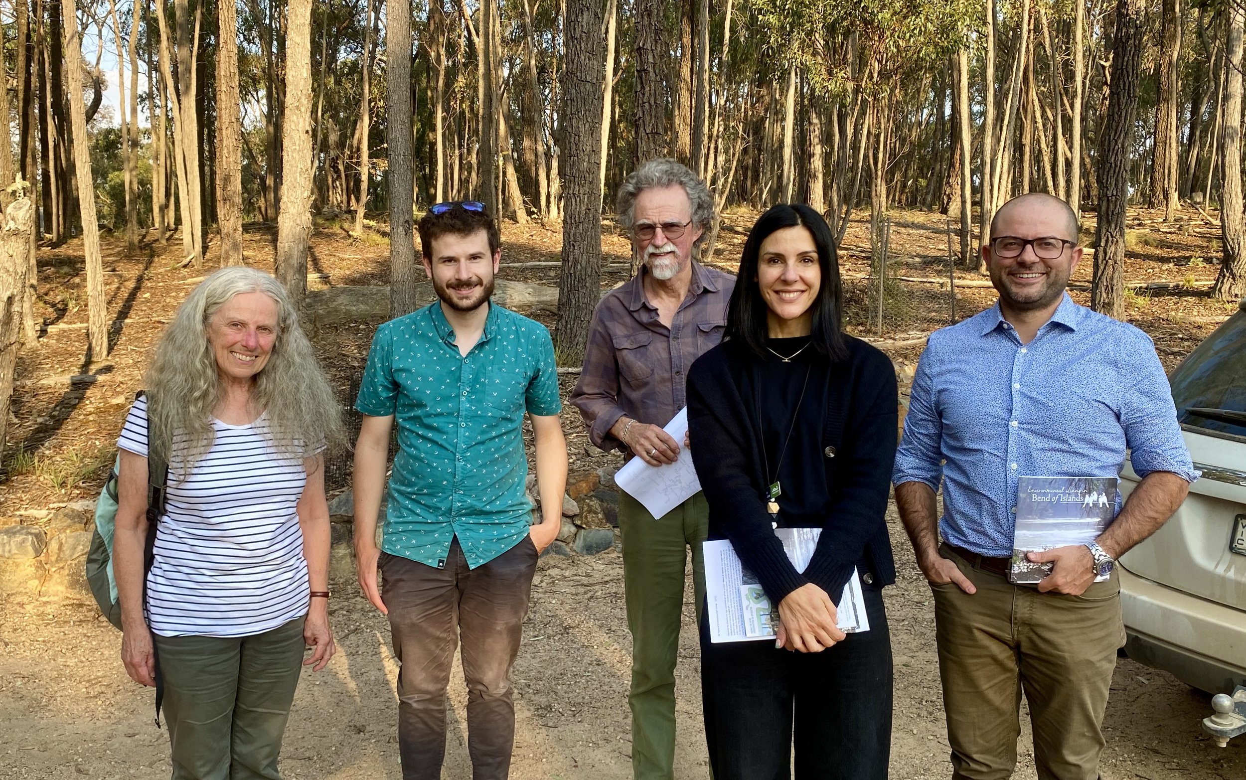 Group of five people standing outdoors in a wooded area, smiling at the camera.