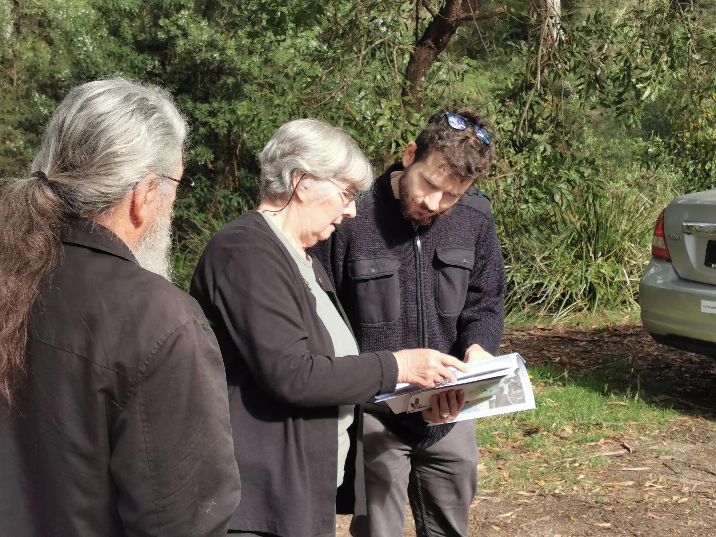 Three people outdoors reviewing a manual or map. Two women and one man are focused on the document, with a car visible in the background.