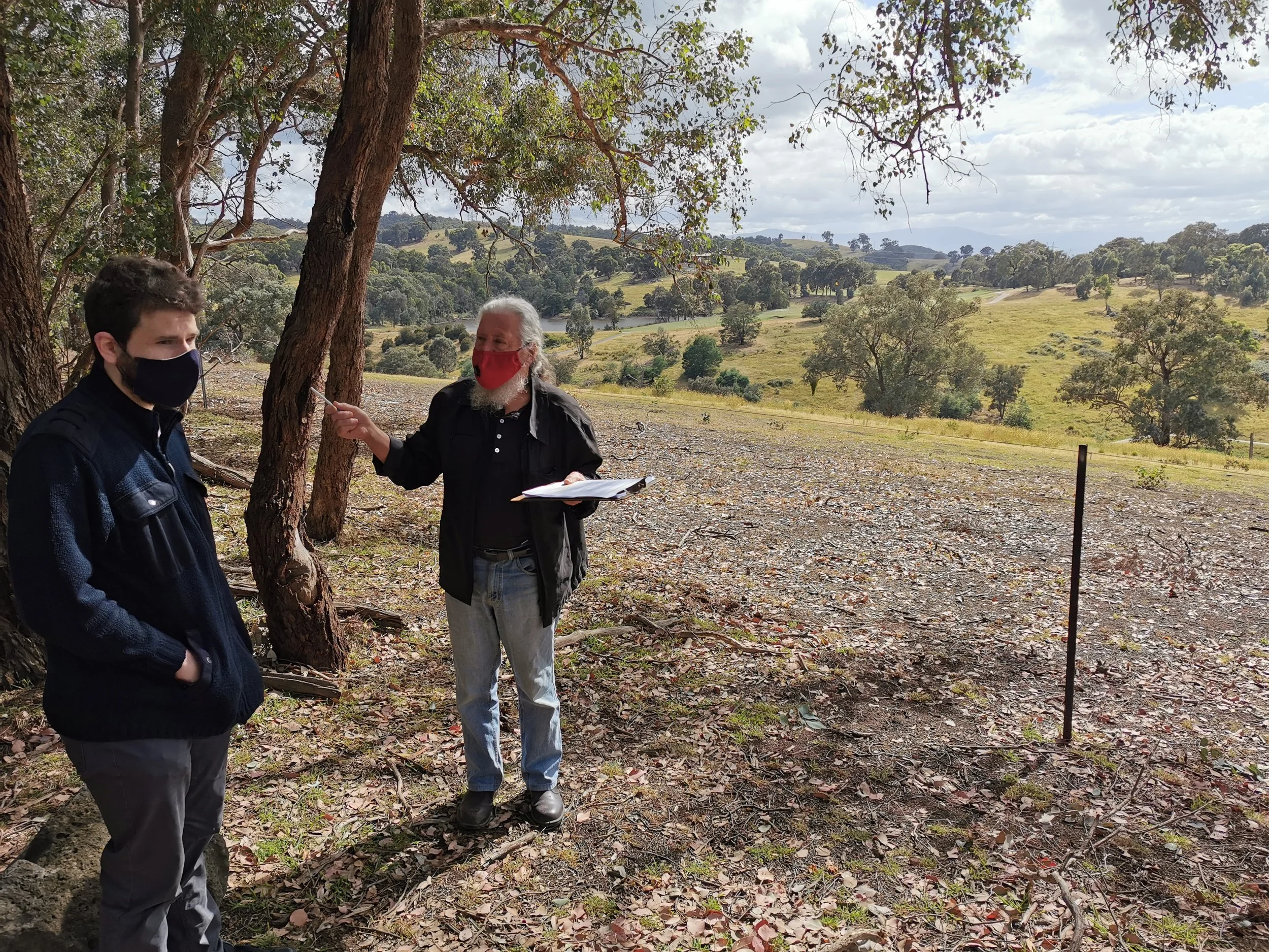 Two men wearing face masks and standing outdoors near trees. One man appears to be speaking and holding a clipboard, while the other listens. The background shows rolling hills and scattered trees under partly cloudy skies.