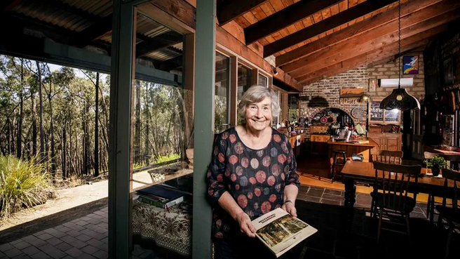 A smiling elderly woman with gray hair holding an open book indoors, standing near a glass door with a view of trees outdoors.