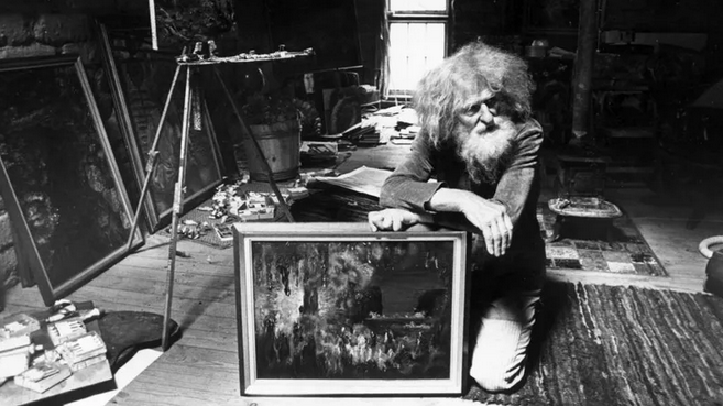 An elderly man with long hair and a beard kneeling next to an artwork in an artist's studio, surrounded by paintings, art supplies, and furniture.