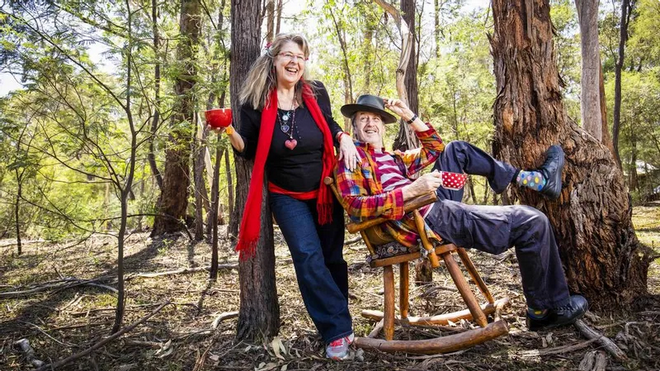 Two smiling women outdoors in a wooded area, one standing and the other sitting in a rocking chair, holding a coffee mug, with trees and sunlight in the background.
