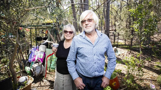 An elderly man and woman standing outdoors in a wooded yard, smiling at the camera. The man has white hair and glasses, wearing a blue shirt, and the woman has short gray hair, wearing sunglasses and a black top.