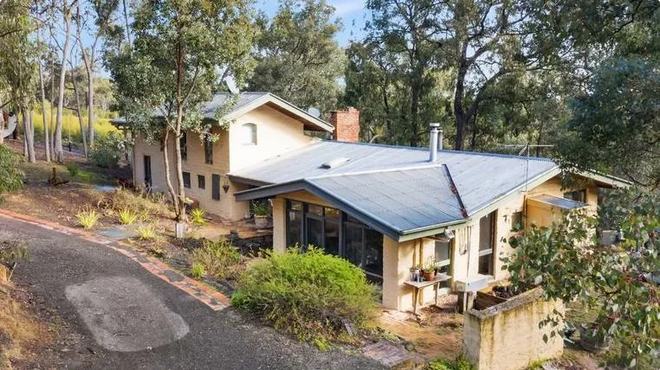 A house in a wooded area with a sloped metal roof, surrounded by trees and a landscaped yard.