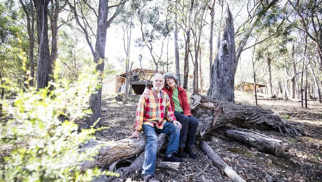 Two people sitting on a fallen tree in a forest, smiling at the camera with trees and a wooden building in the background.