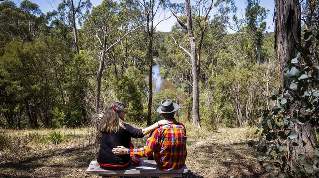 Two people sitting on a bench outdoors, overlooking a river surrounded by trees.