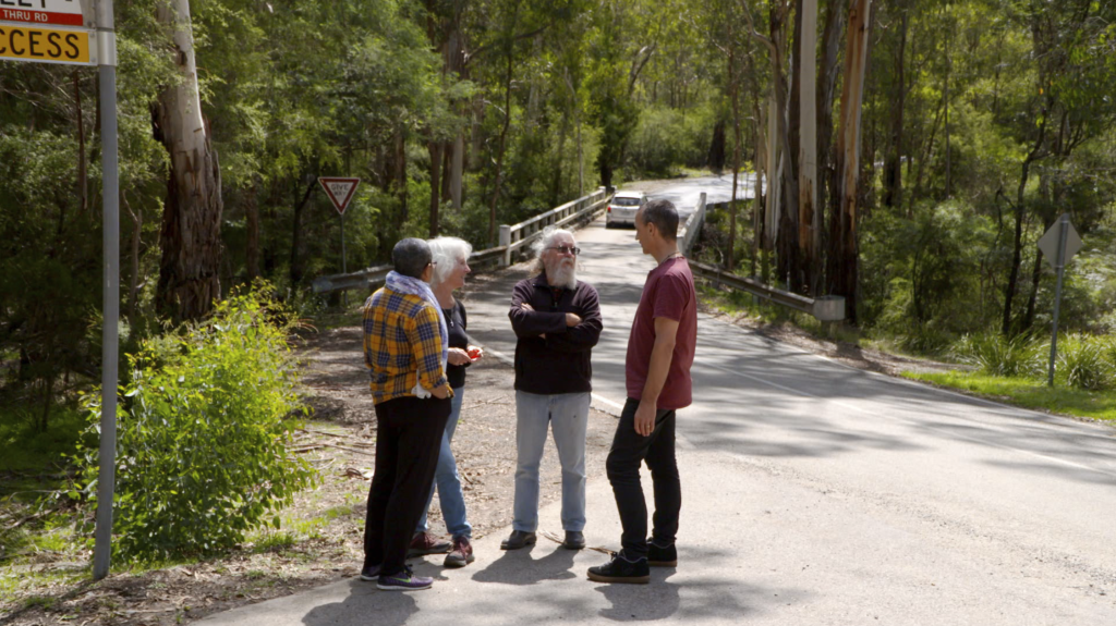 Four people standing and talking on a rural road surrounded by trees, with a bridge in the background and various road signs visible.