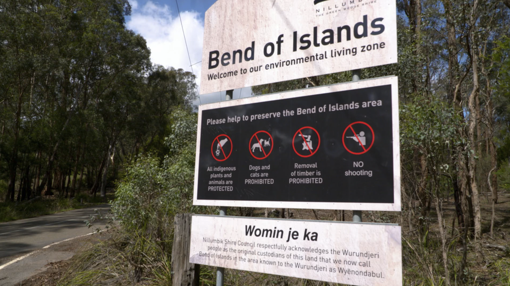 A signboard at Bend of Islands area with instructions to protect local flora and fauna, prohibiting dogs, cats, timber removal, and shooting, amidst trees and a road.