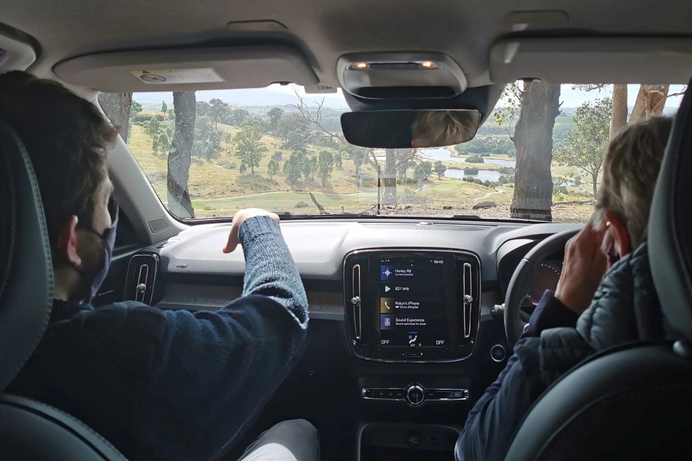 The interior of a vehicle with two passengers driving through a scenic landscape with trees and hills visible through the windshield.