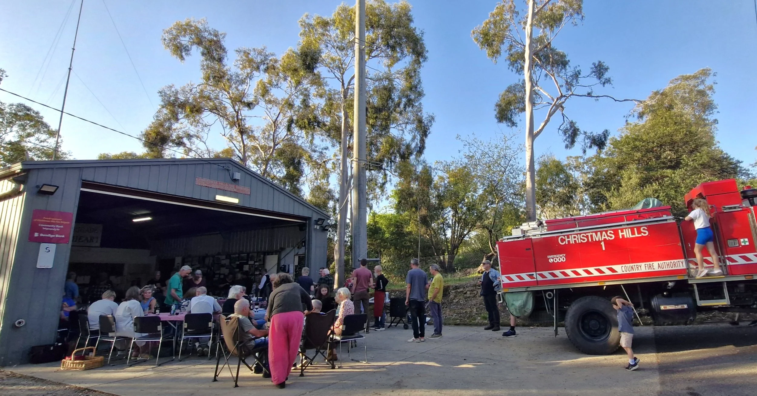A gathering of people inside and outside a CFA fire station shed; some are seated, others standing, with gum trees in the background.