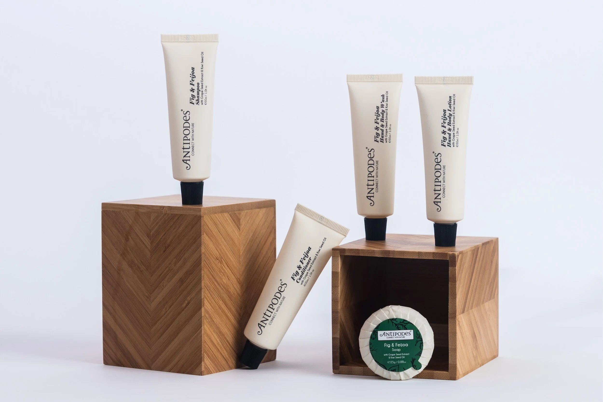 A photo of ANTIPODES Fig and Feijoa Shampoo, Conditioner, Body Wash, Body Lotion and a soap bar are perched artistically on herringbone patterned wooden plinths. They are set against a white background.
