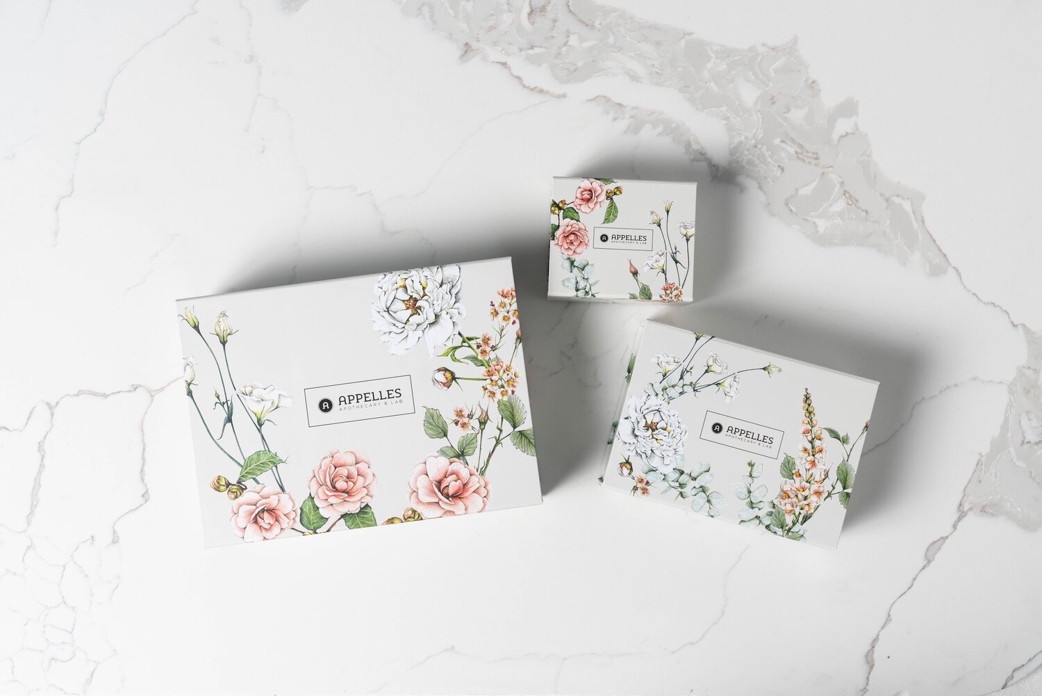 A photo of 3 boxes on a marble table. The boxes are 3 different sizes, small, medium and large. There are scattered flowers adorned on them, with APPELLES brand.