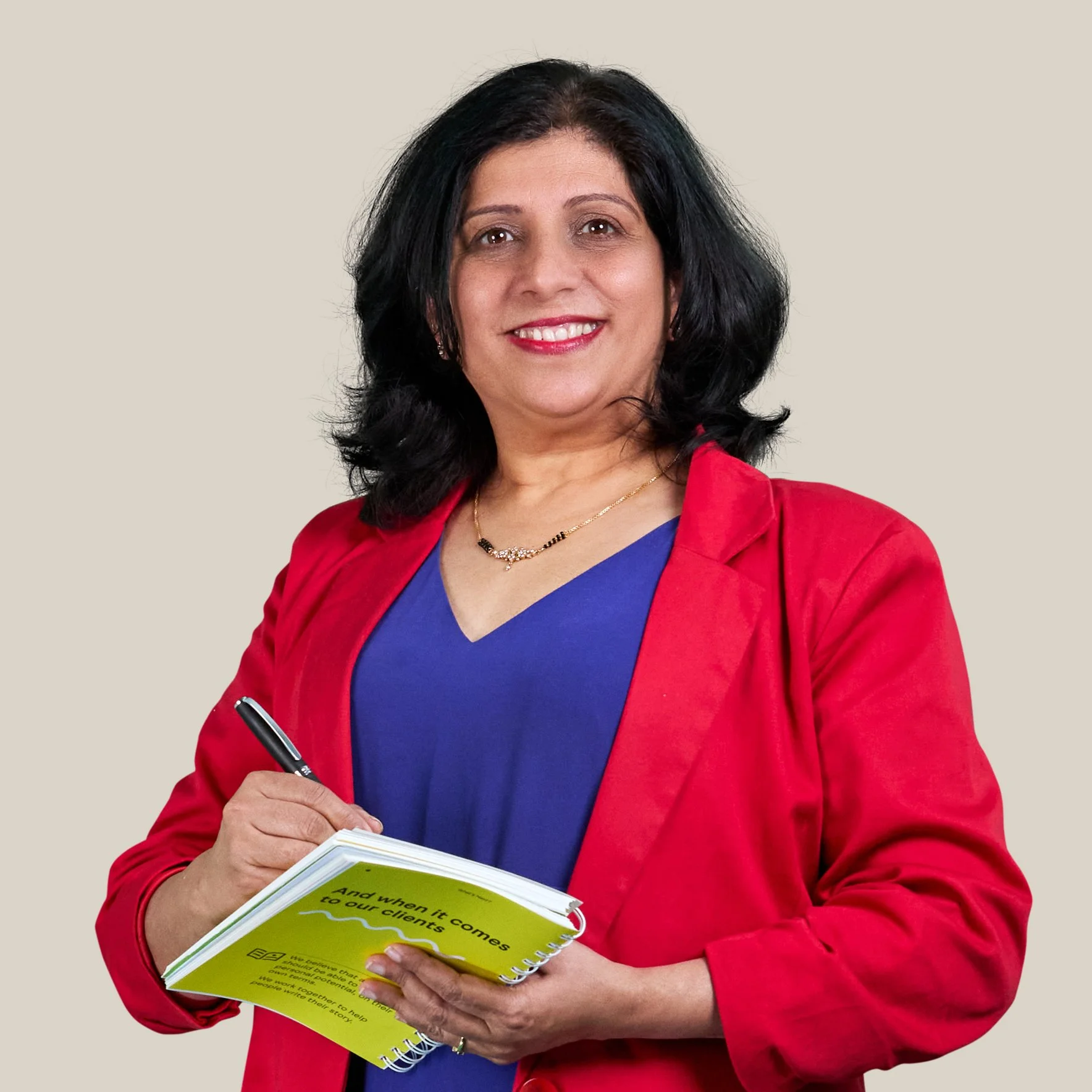 A woman is standing against a grey background wearing a red blazer and purple top. She holds a notepad and a pen ready to write in it while looking at the camera.