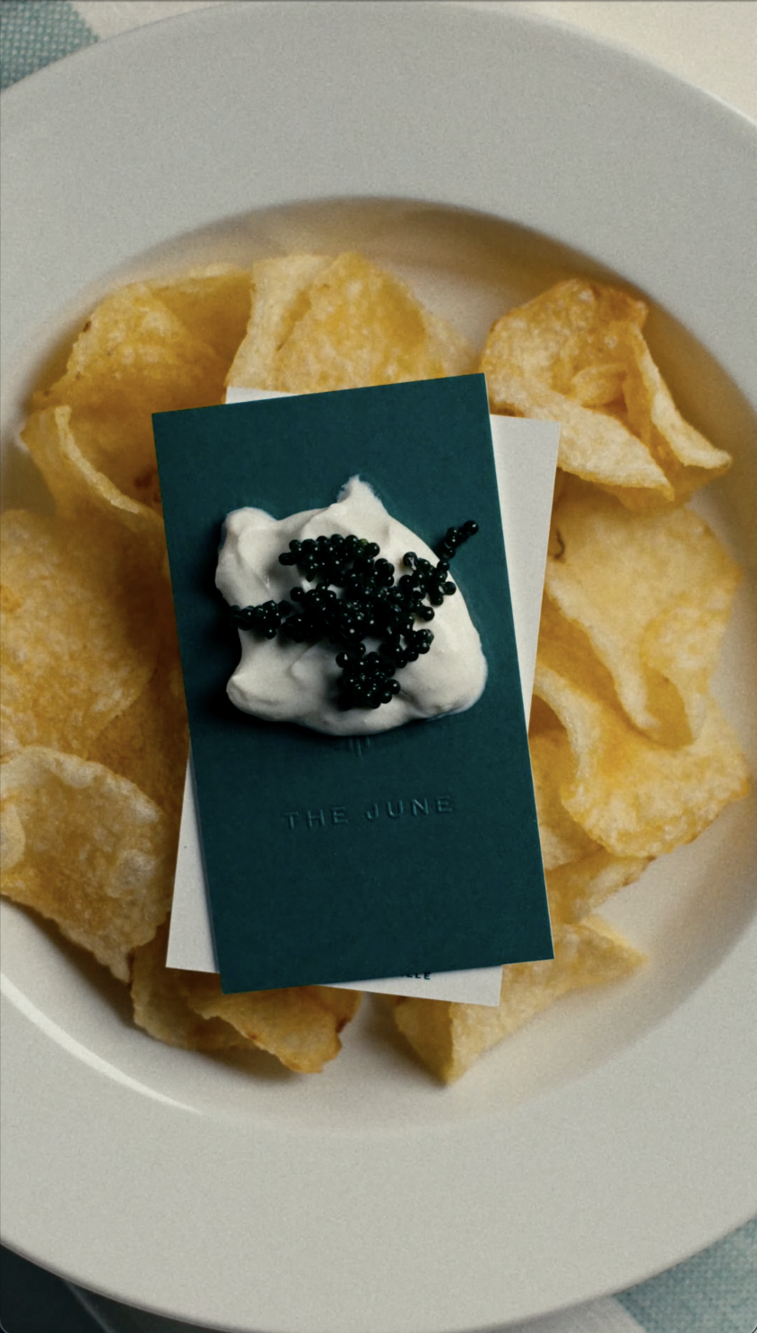 Potato chips on a white plate with a dark green card on top that has whipped cream and black caviar, and the words 'The June' printed on it.