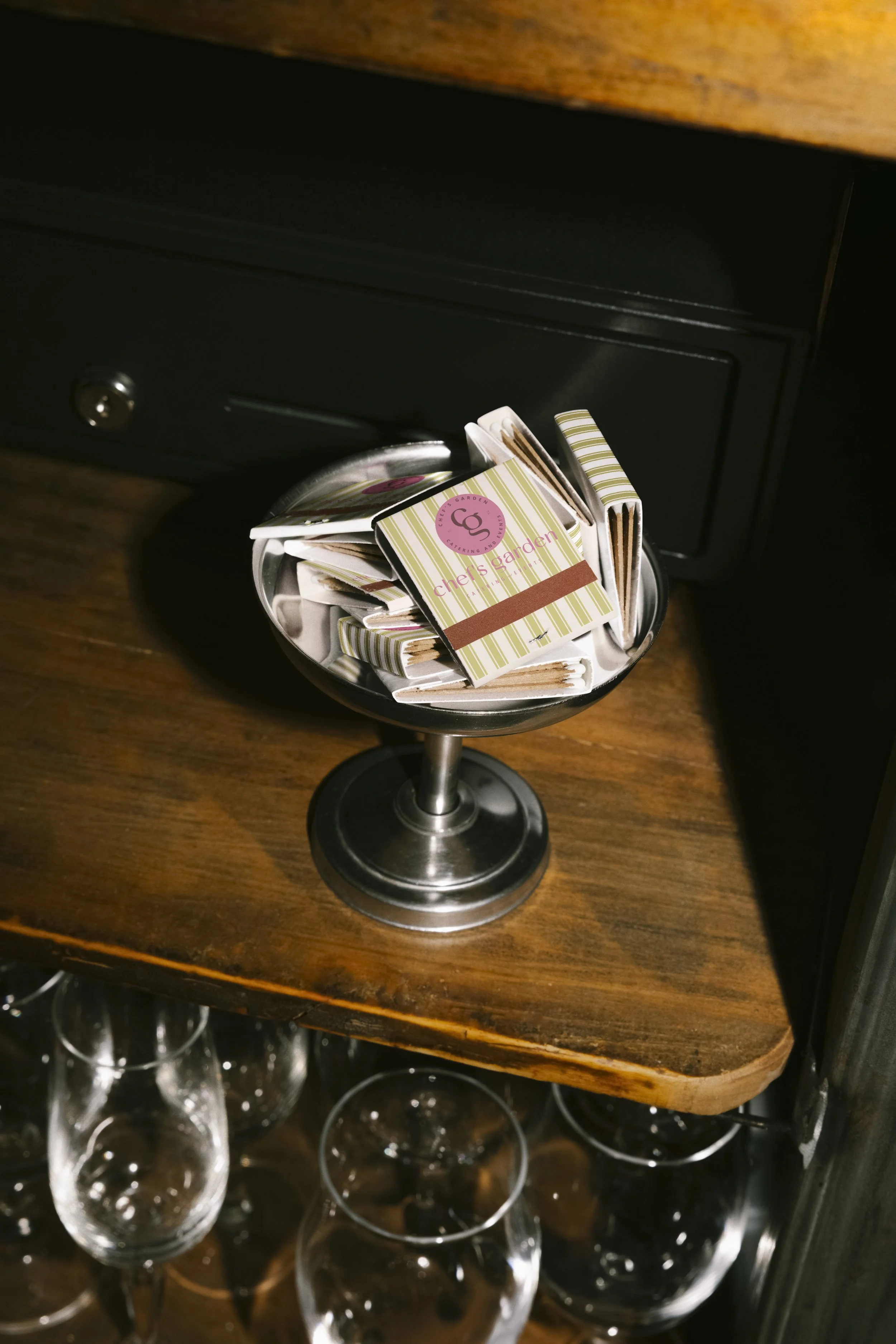 A metal bowl filled with matchbooks on a wooden shelf near glassware underneath.