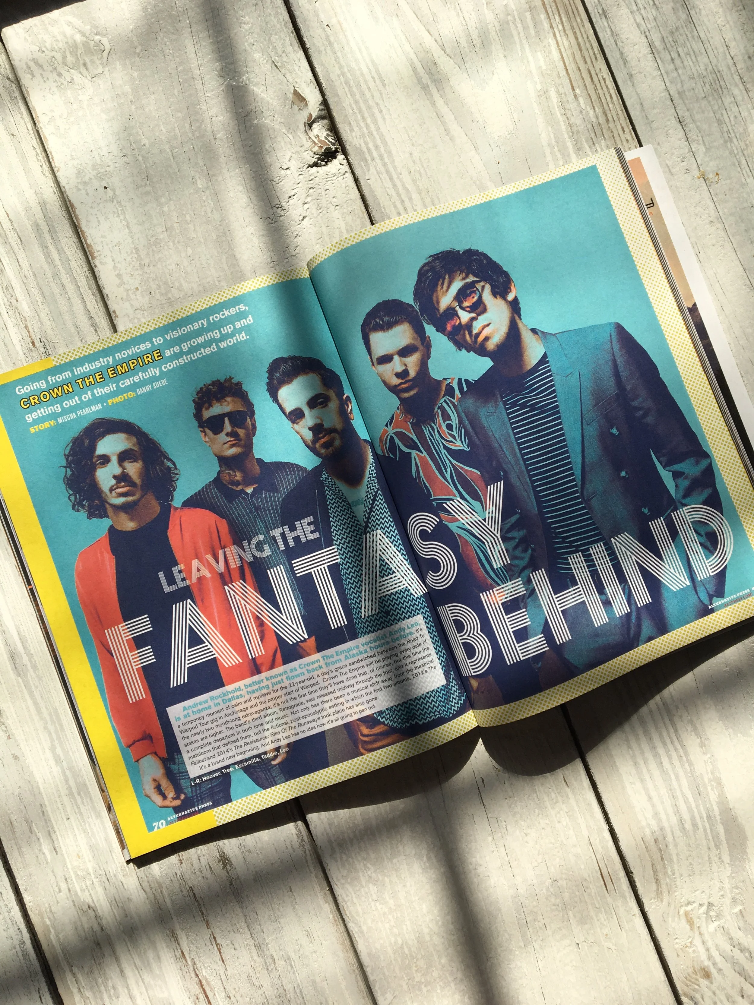 Open magazine on a wooden surface showing a two-page spread featuring the band Crown the Empire with the heading "Leaving the Fantasy Behind" and a group photo of five band members.
