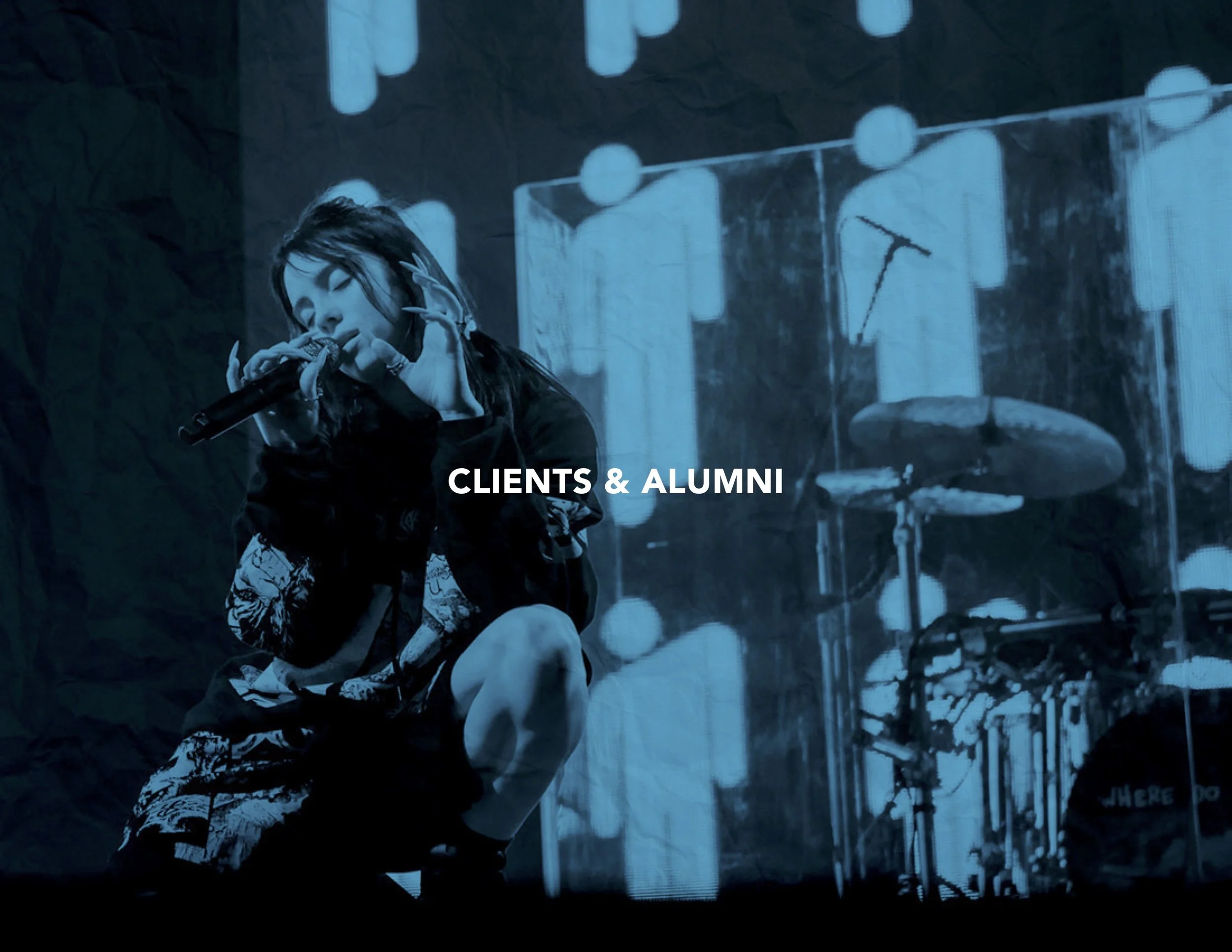 Billie Eilish singing into a microphone on stage with a dim background and drums.