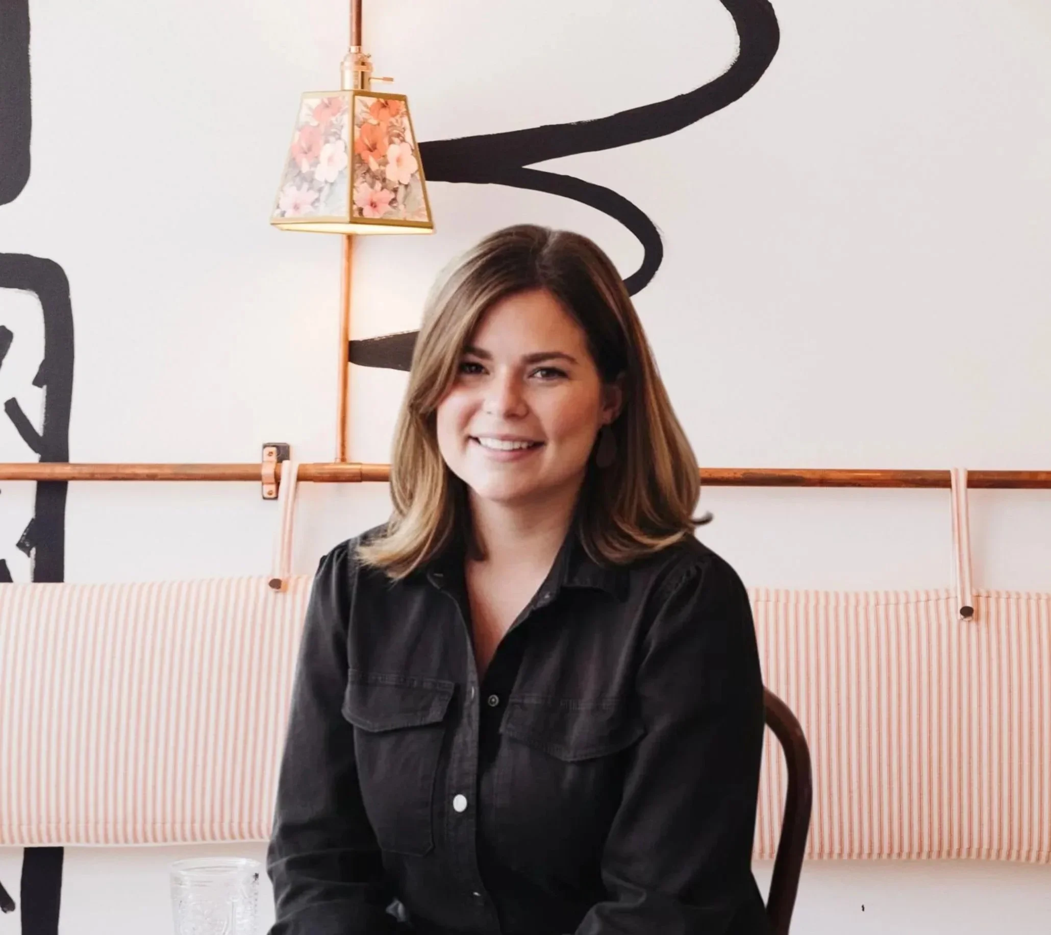 A smiling woman with shoulder-length brown hair seated indoors with a striped cushion behind her and a wall with black line art and a floral lamp in the background.