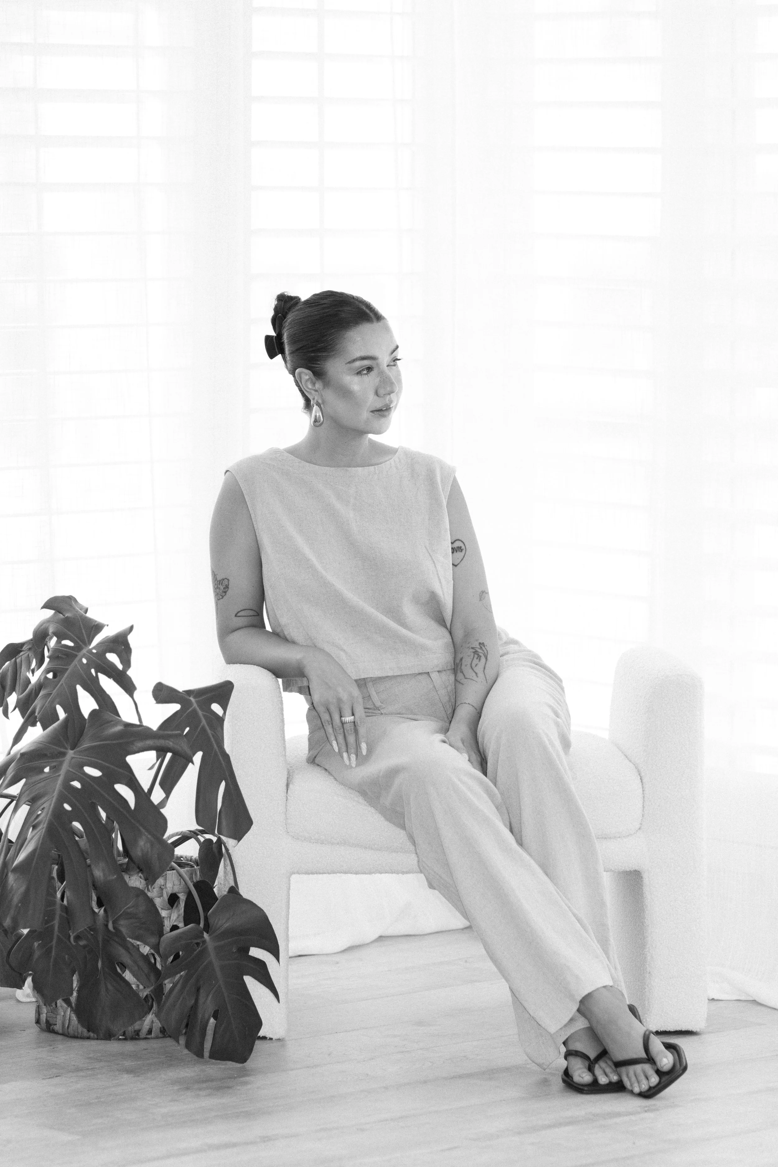 A woman sitting on a white upholstered chair in front of a bright window, with a plant nearby, wearing a sleeveless top, wide-leg pants, and sandals, with hair styled in an updo and tattoos visible on her arms.