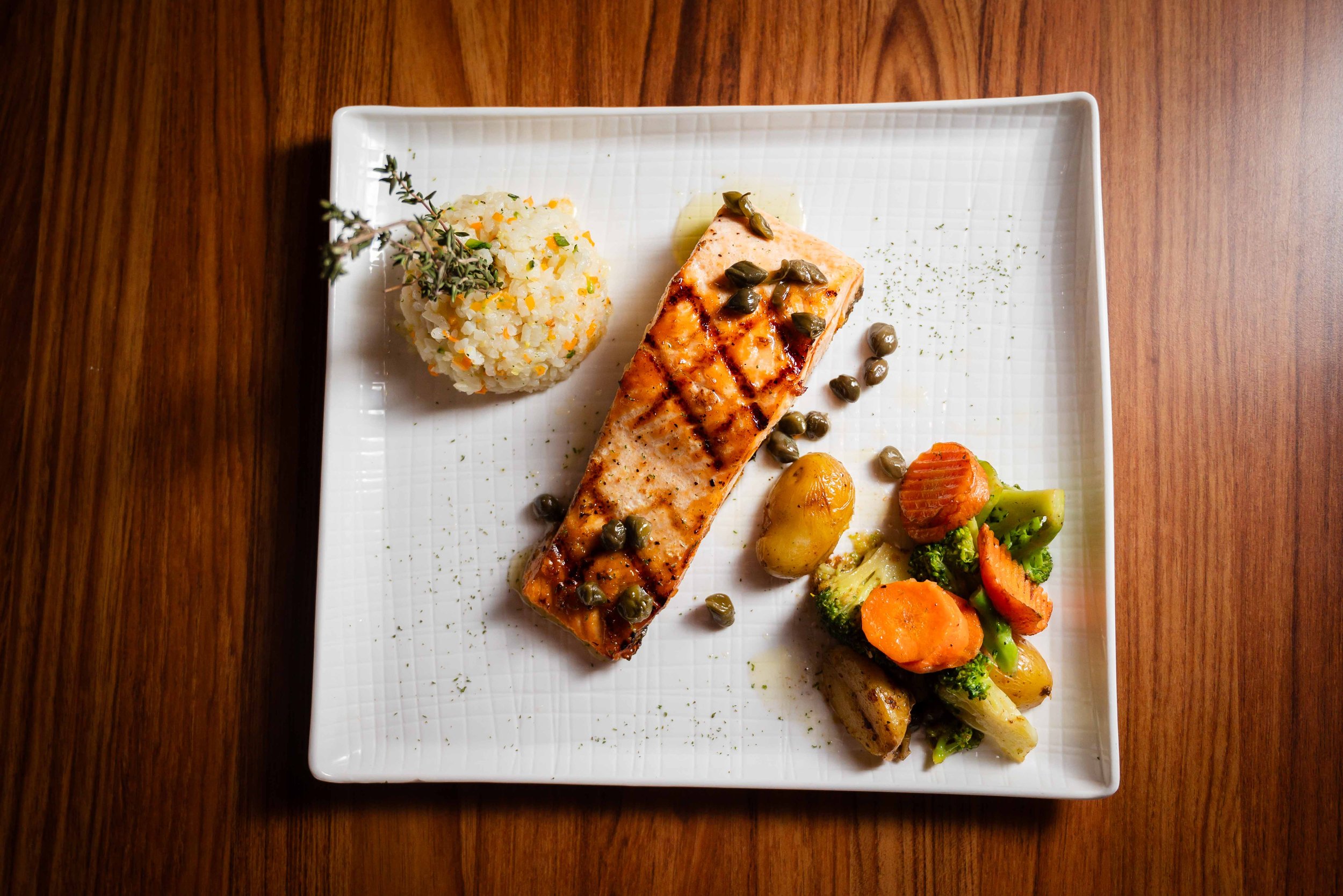 Plato de salmón a la parrilla con alcaparras, acompañado de arroz con hierbas, papas asadas y verduras.