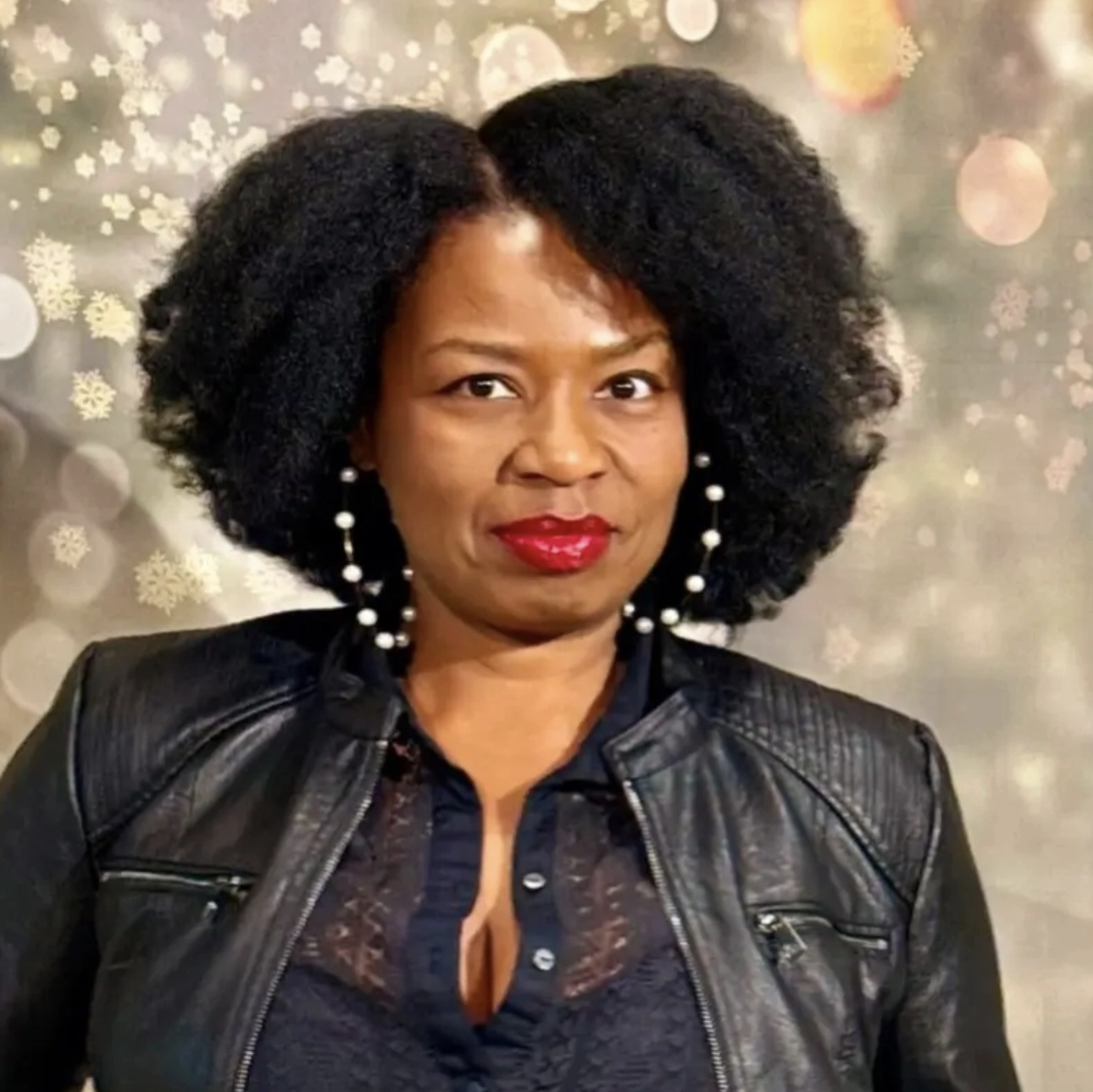 Portrait of Nikei S. Salas. She is pictured in a black leather jacket with a black top and large black and white beaded hoop earrings. Her hair is natural and shoulder length.