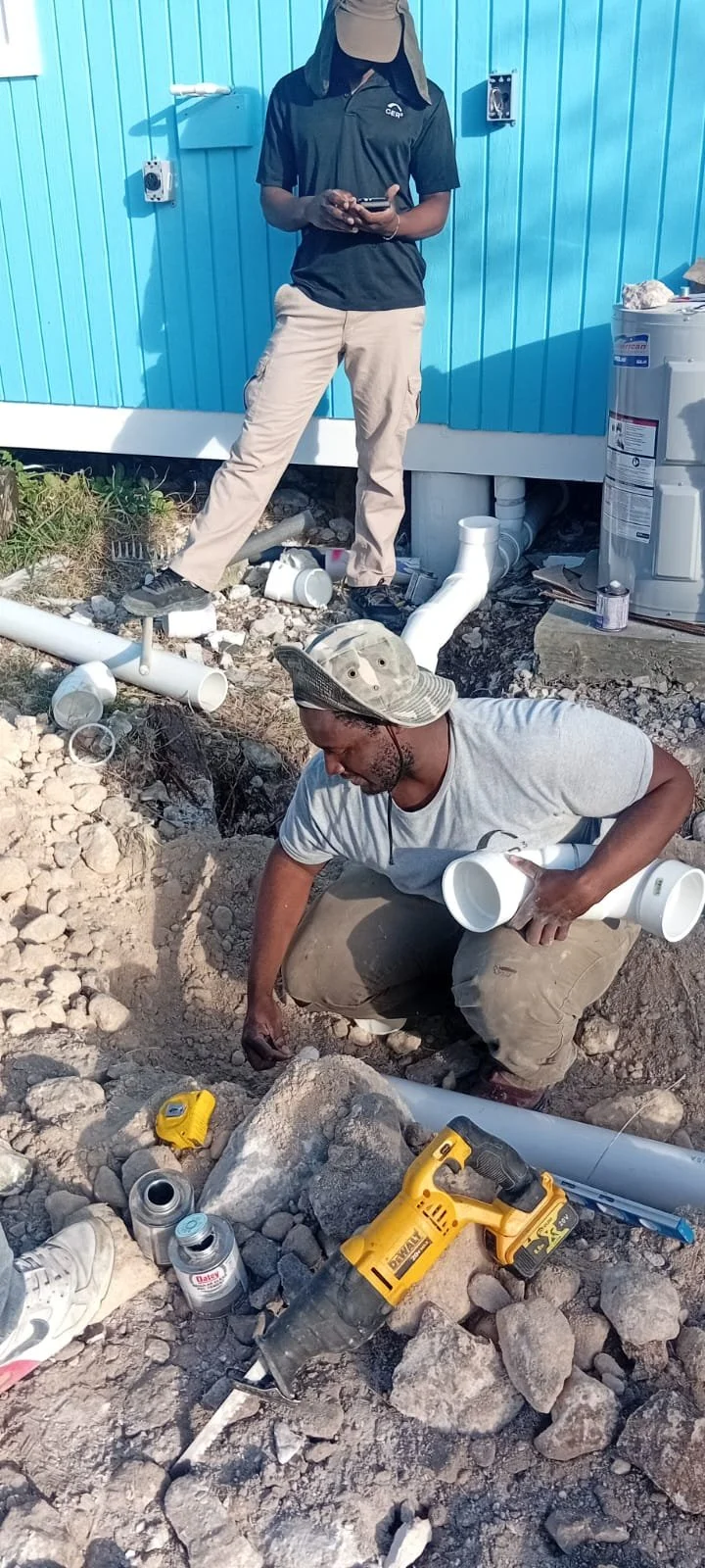 Two men working on a plumbing project outside a blue building. One is standing, wearing a beige hat and black shirt, looking at a device. The other is crouching, wearing a camouflage hat and gray shirt, holding PVC pipes, with tools and pipes around