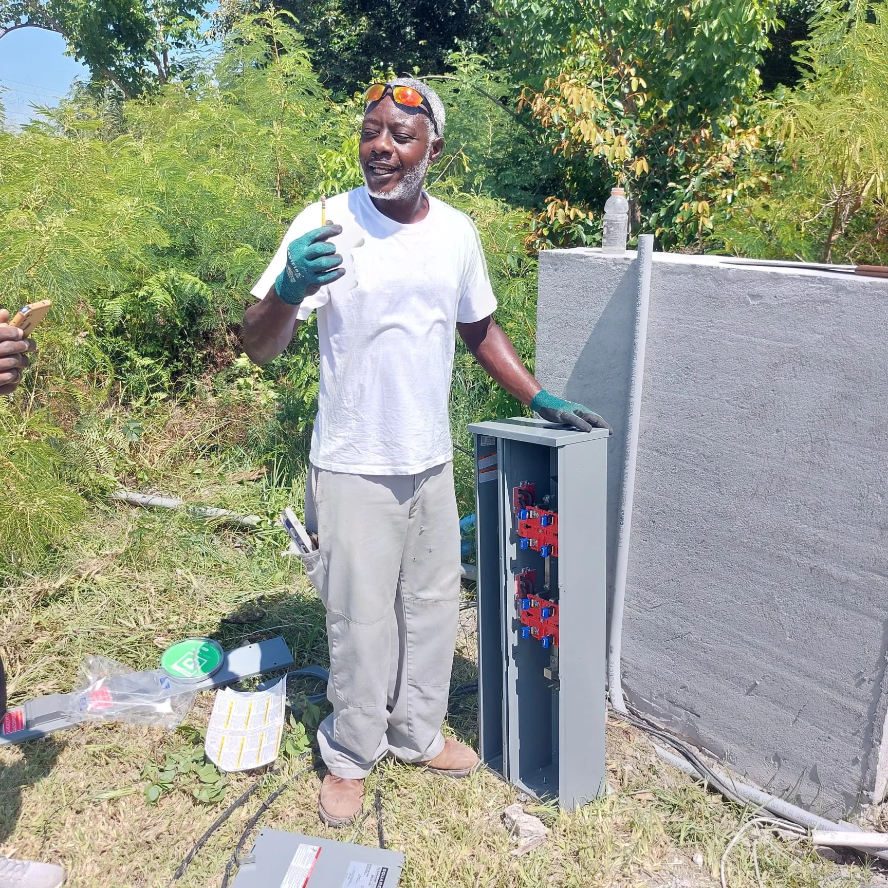 A man working on an outdoor electrical panel surrounded by greenery, wearing gloves and holding a tool, with electrical equipment and documents nearby.