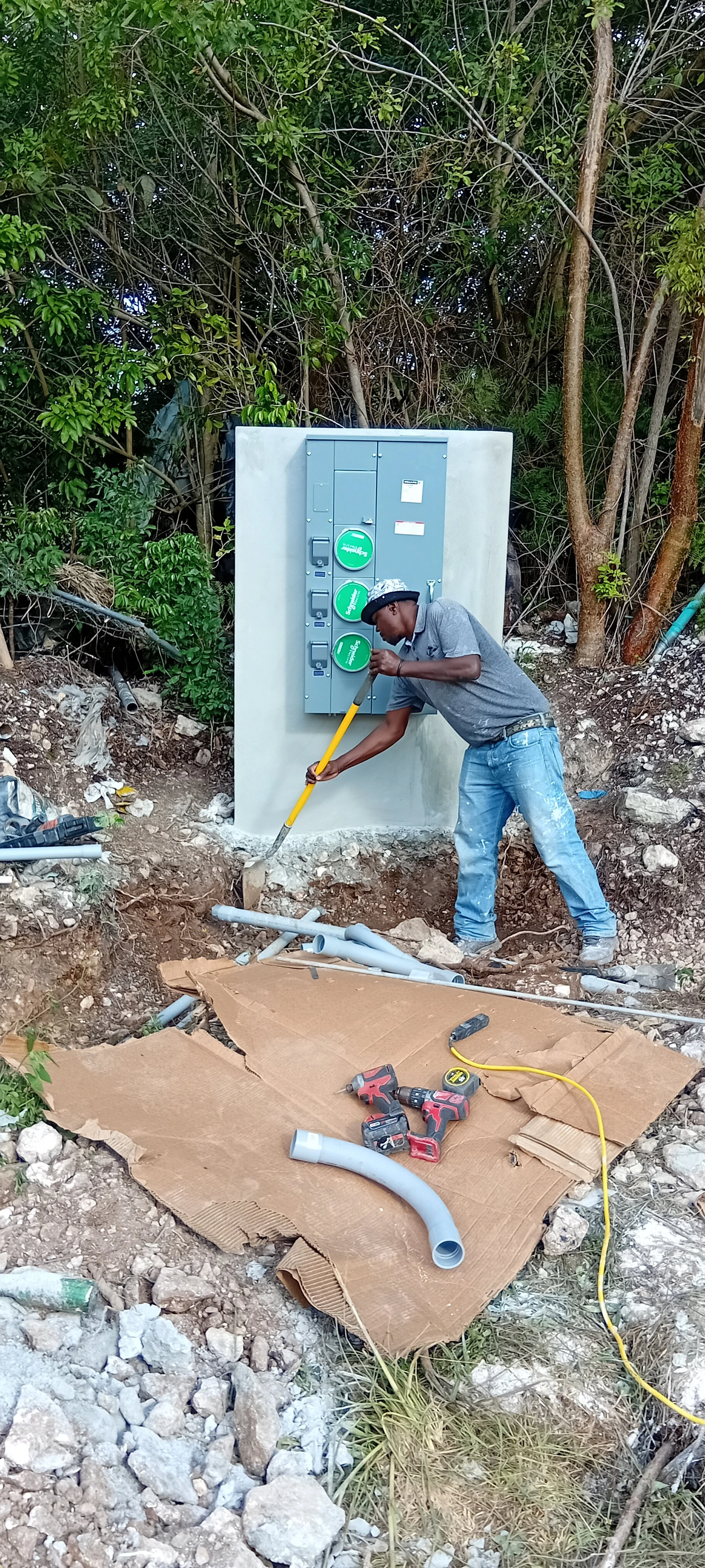Man working on electrical or utility box installation outdoors, surrounded by trees and construction tools, with peeled pipes on the ground.