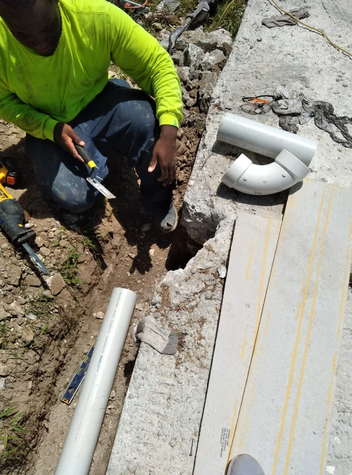 A construction worker in a neon yellow shirt working with PVC pipes and fittings outdoors. His tools include a utility knife, and the site has concrete, rocks, and other construction materials.