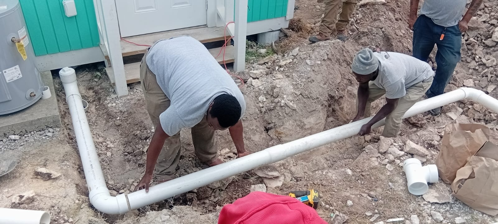Two workers installing white PVC pipes in a trench outside a house under construction.