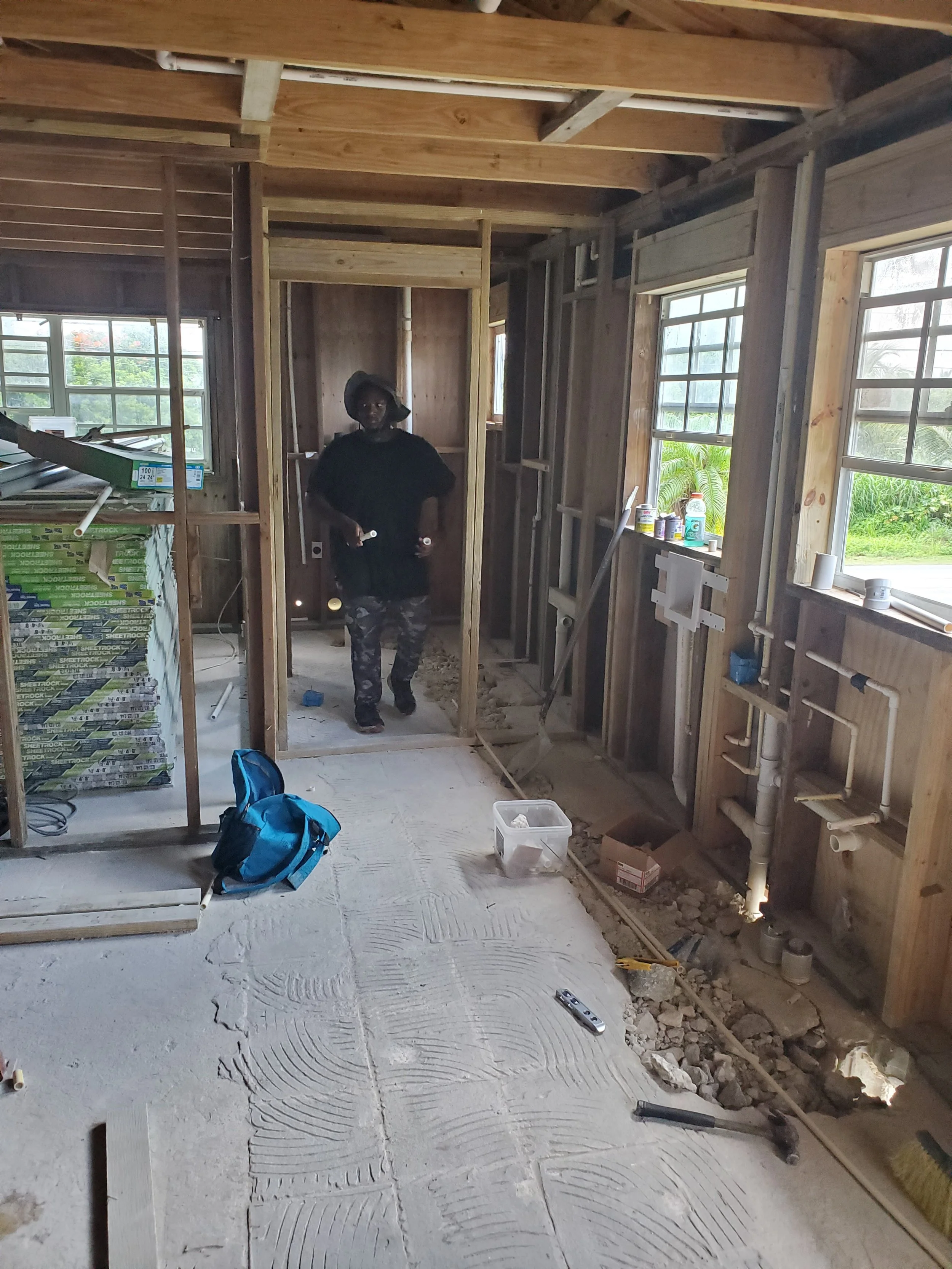 A person standing inside a house under construction, with exposed wooden framing, construction tools and supplies, and a partially finished cement floor.