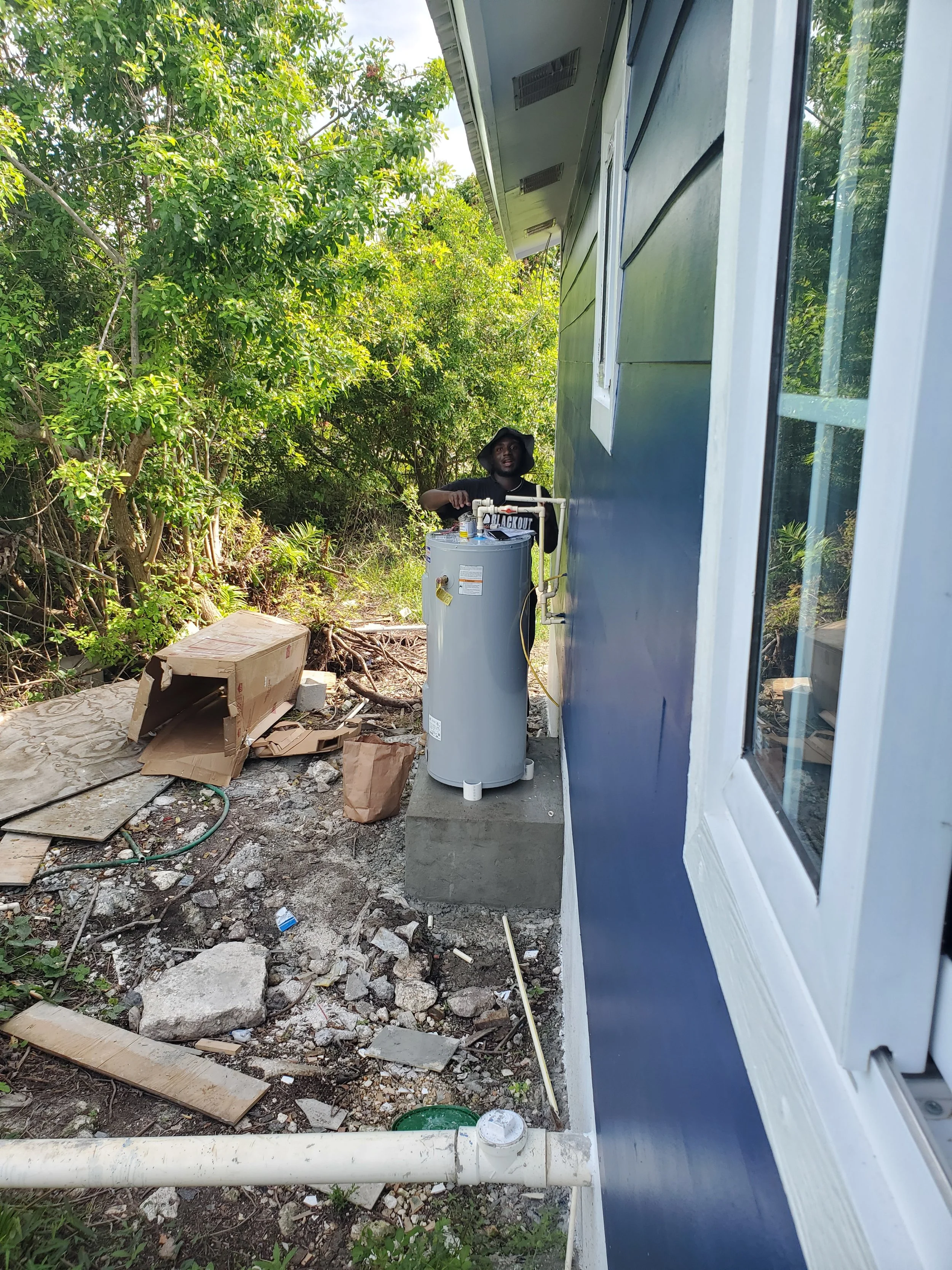 Person installing or repairing a water heater outside a house with blue siding, surrounded by trees and construction debris.