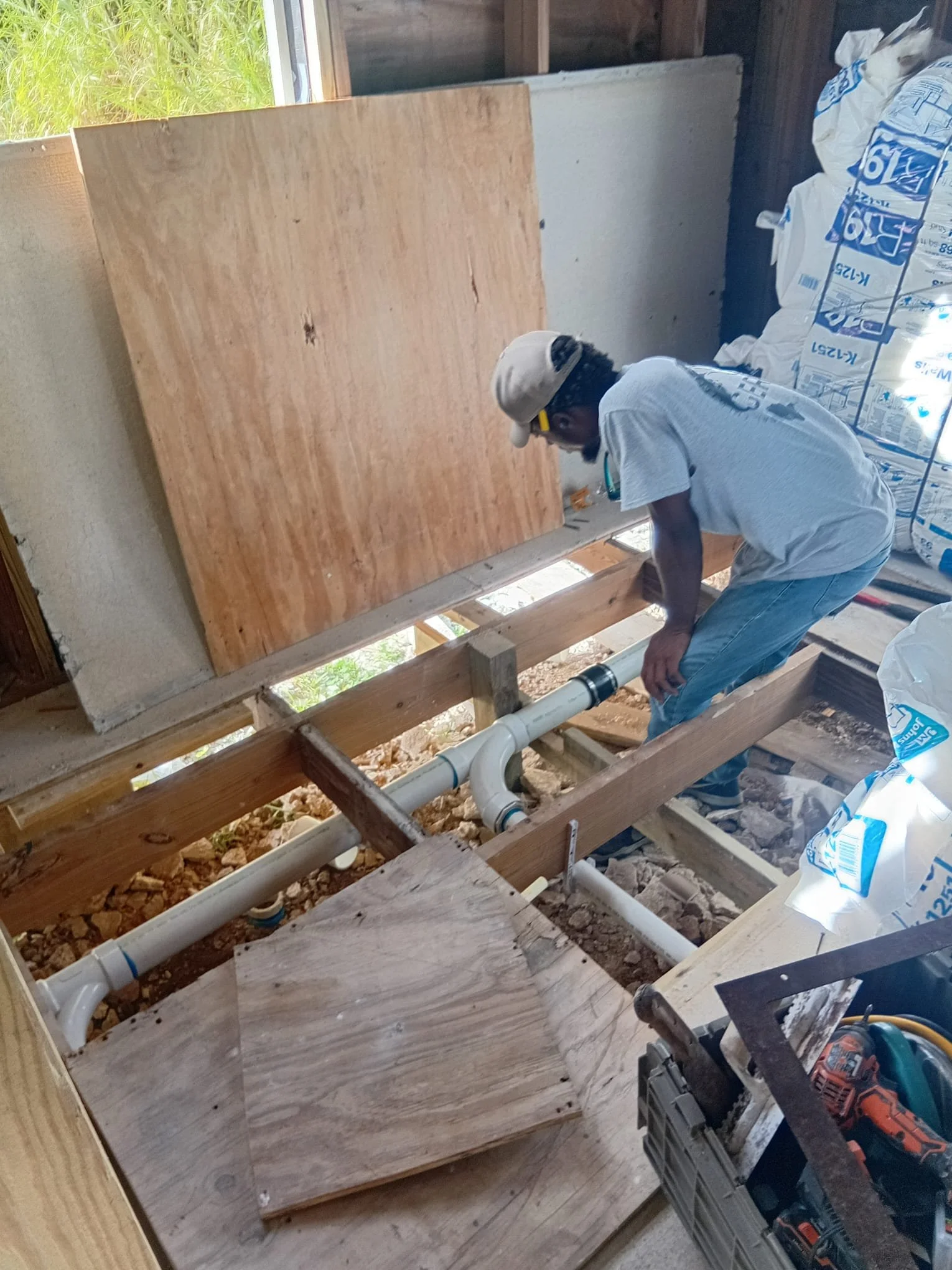 A person installing plumbing pipes inside a wooden floor in a construction site. The worker is wearing a cap, glasses, and safety goggles.