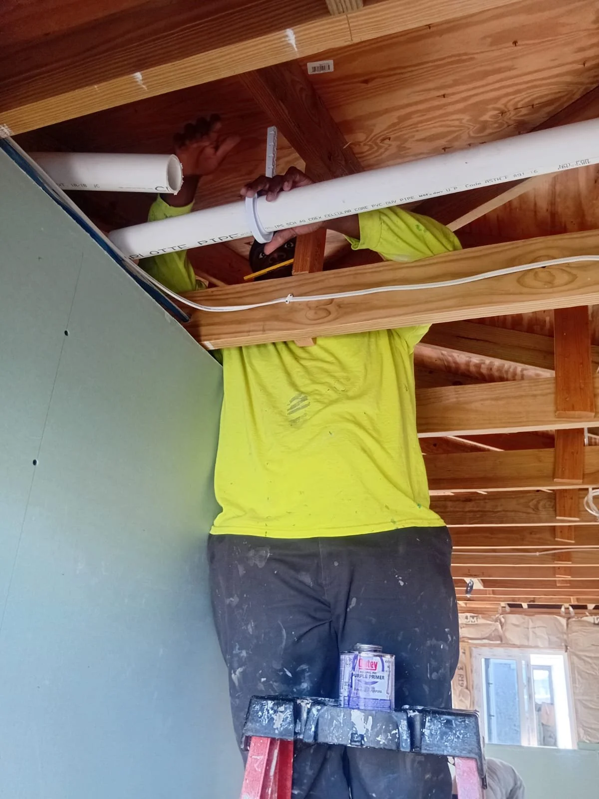Construction worker installing or assembling a white PVC pipe in a building under construction, standing on a ladder with a can of primer and drywall in the background.
