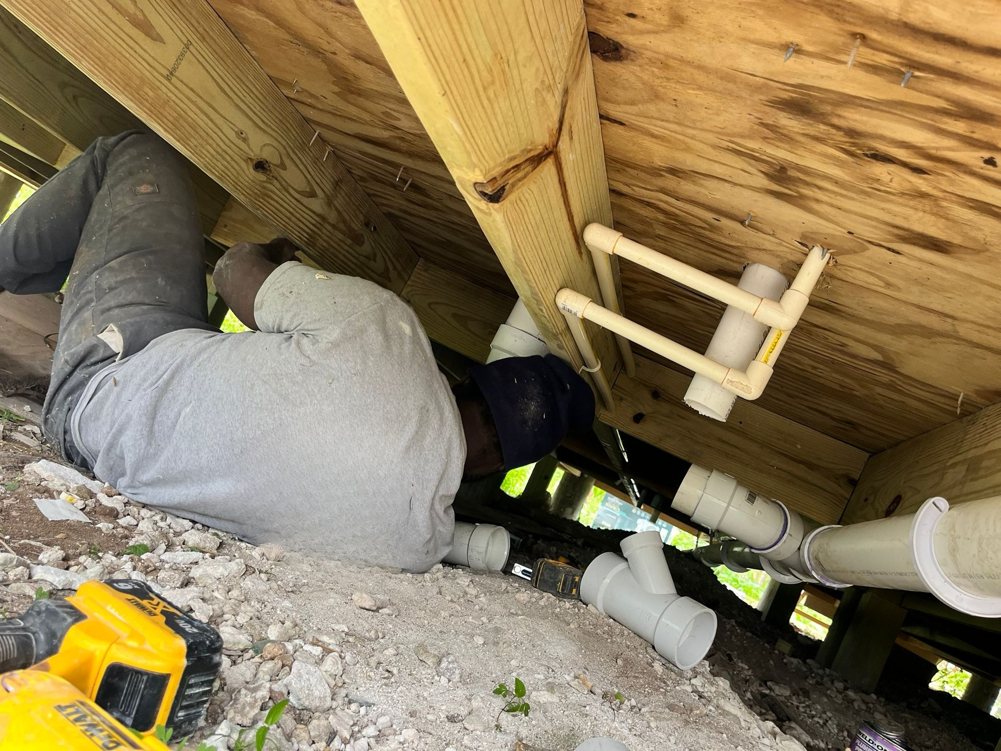 Two workers installing plumbing pipes under a wooden structure, with tools and gravel on the ground.