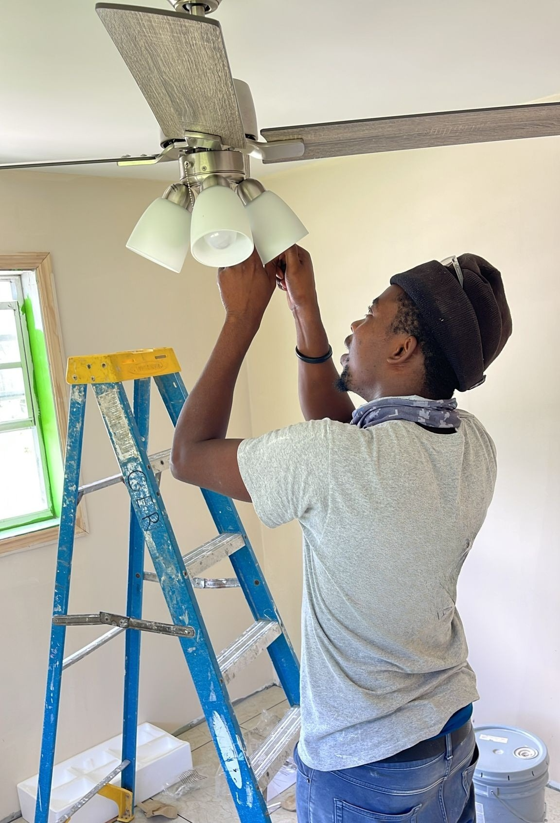 A man wearing a gray t-shirt, blue jeans, and a black beanie is standing on a blue ladder, adjusting a ceiling fan with four lights in a room with pale walls and a window.