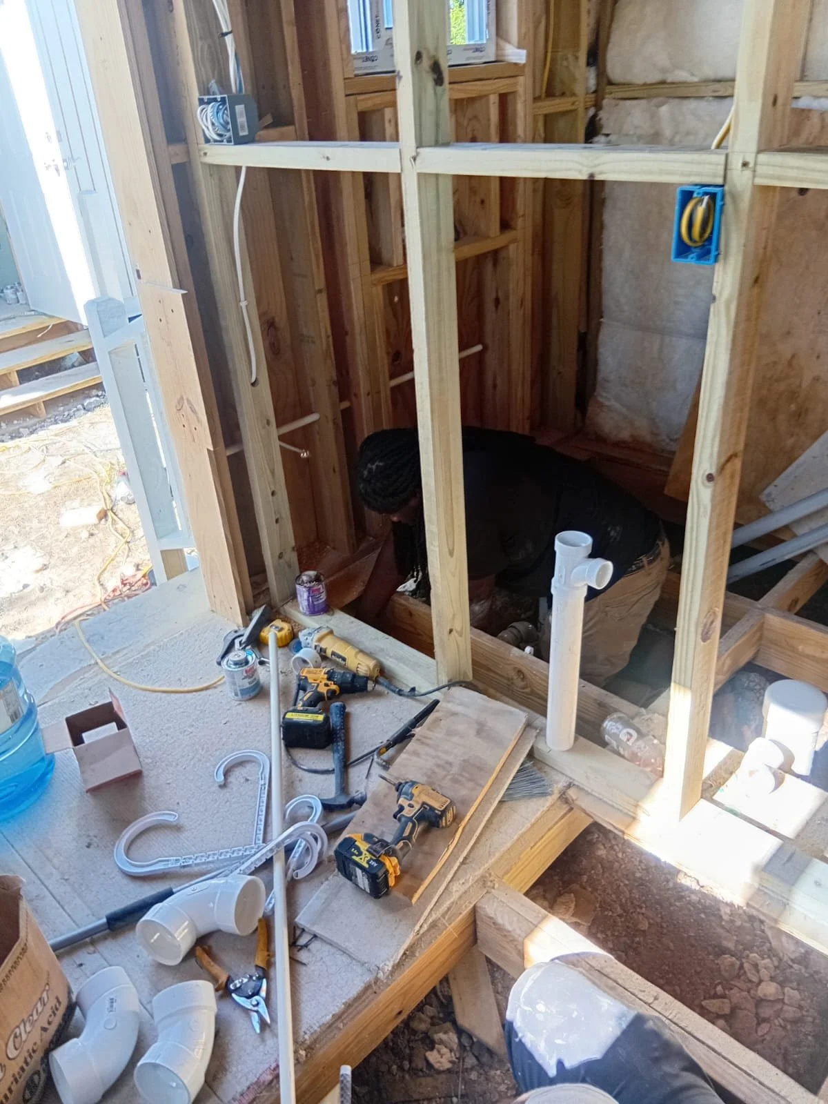 A person working on plumbing inside a partially framed house with construction tools and pipes on the floor.