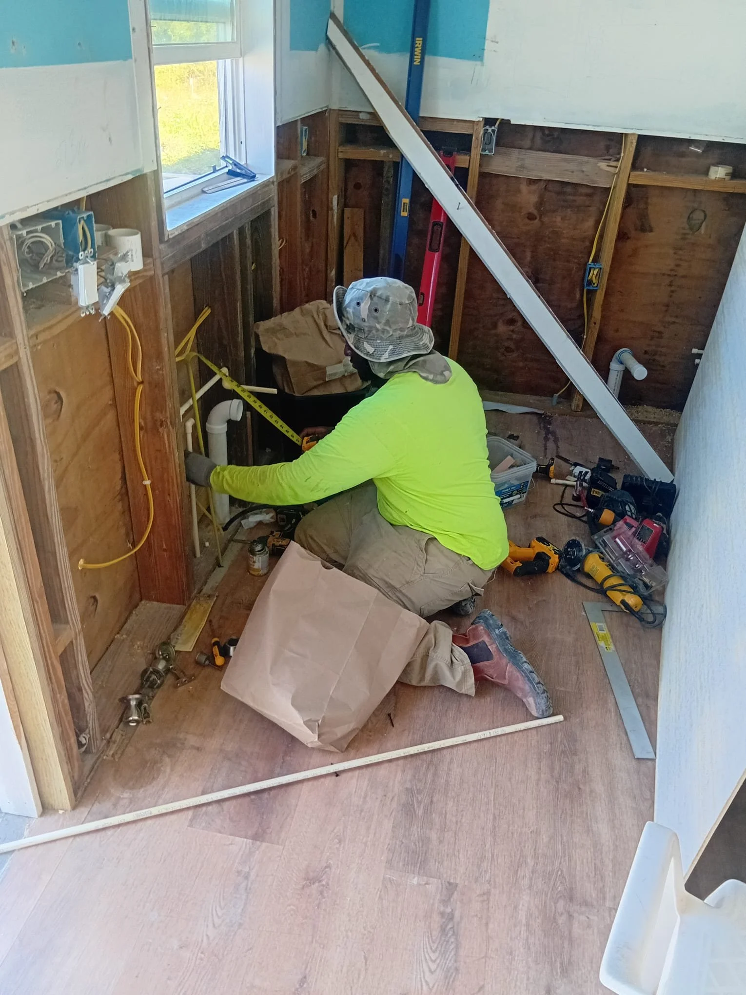 A worker in a bright yellow shirt and a camouflage hat installing plumbing in a partially constructed room, surrounded by tools and construction materials.