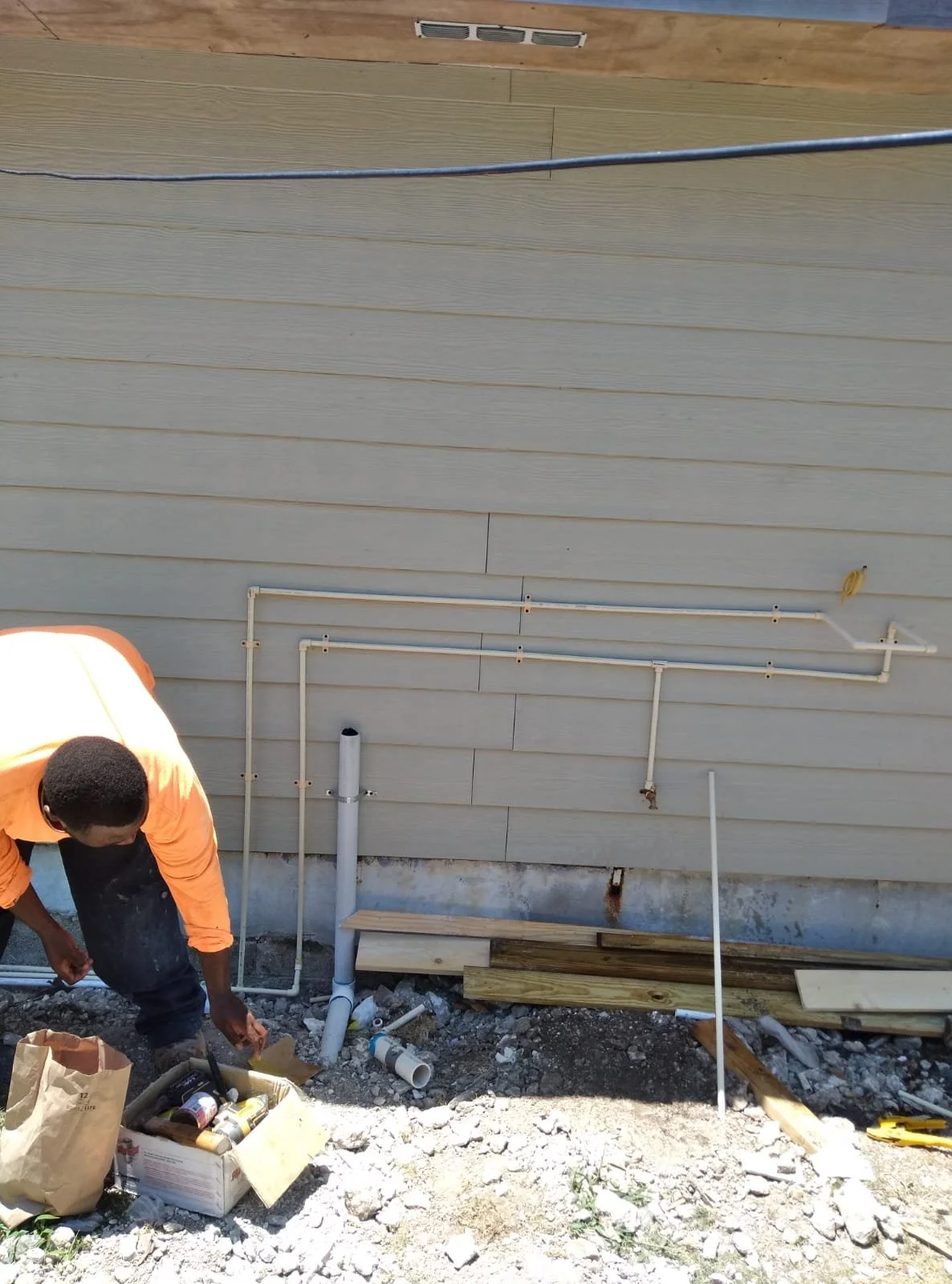 A person working on an outdoor plumbing installation with PVC pipes on the side of a house, surrounded by construction debris.