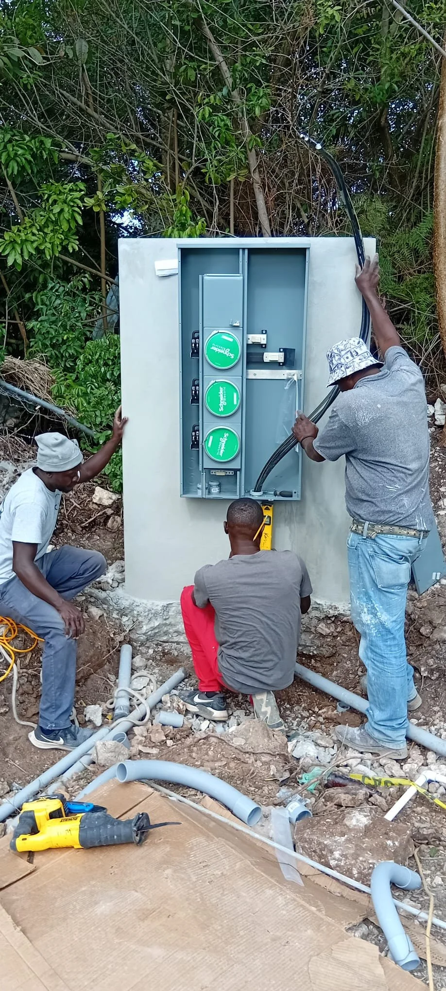 Three workers installing or repairing a utility box outdoors among trees, with tools and pipe fittings on the ground.