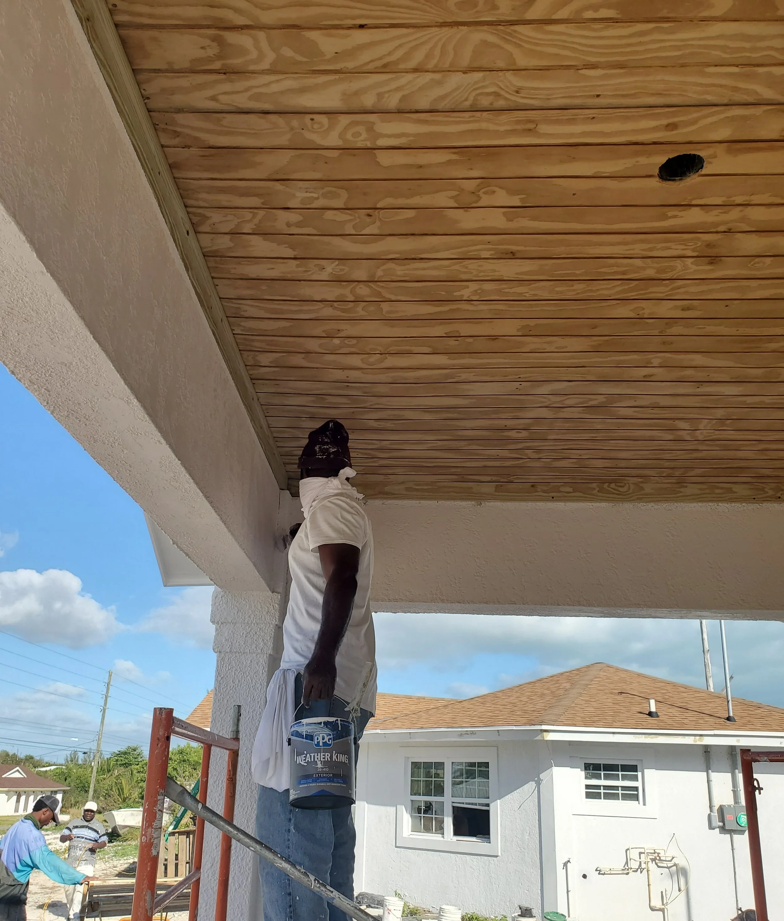 A man standing on a scaffold painting the underside of a house's roof overhang while holding a can of paint, with other workers visible in the background during daytime.
