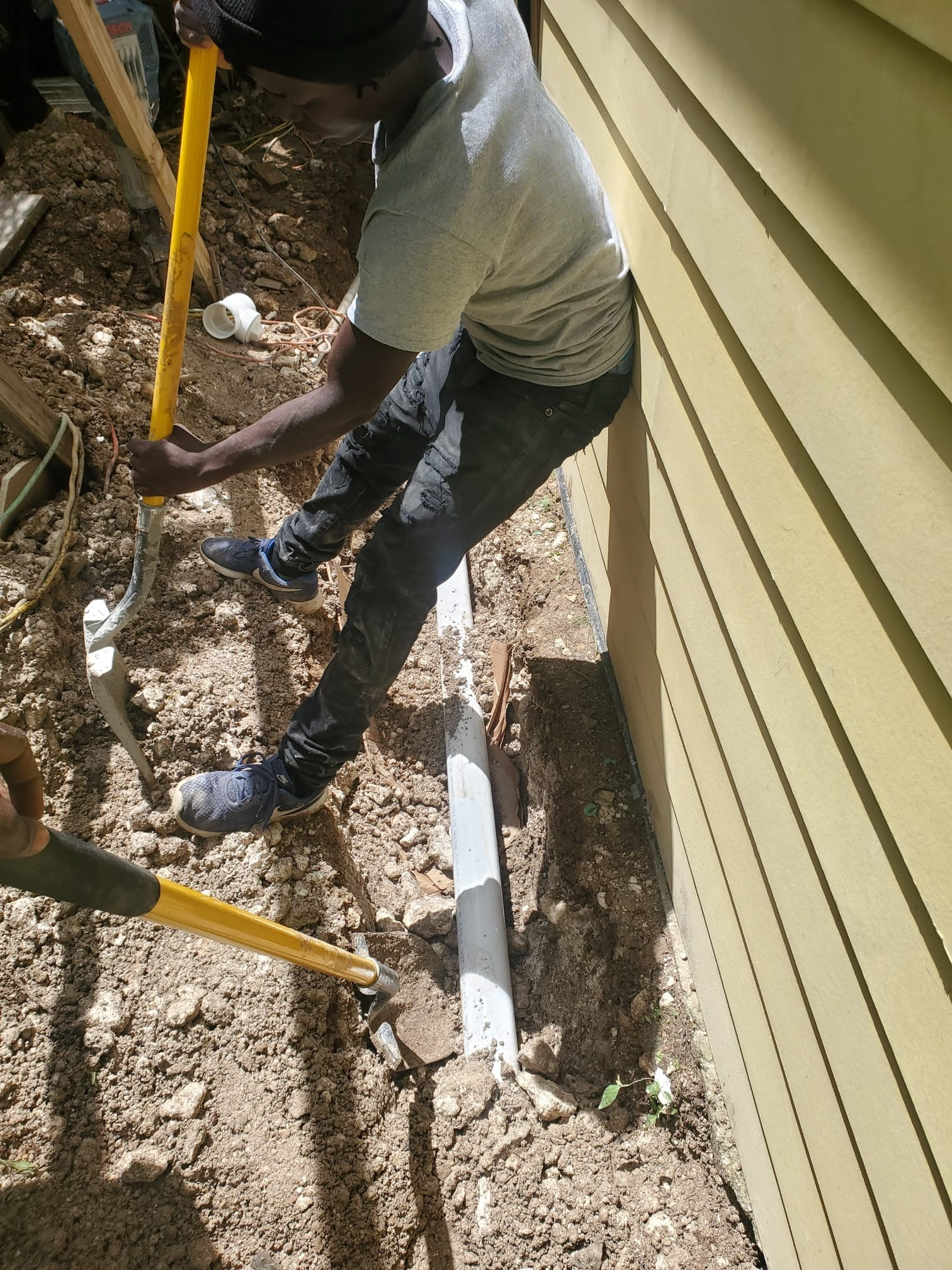 A person working outside next to a yellow house, digging in the dirt with a yellow-handled shovel, near a white pipe along the house foundation.