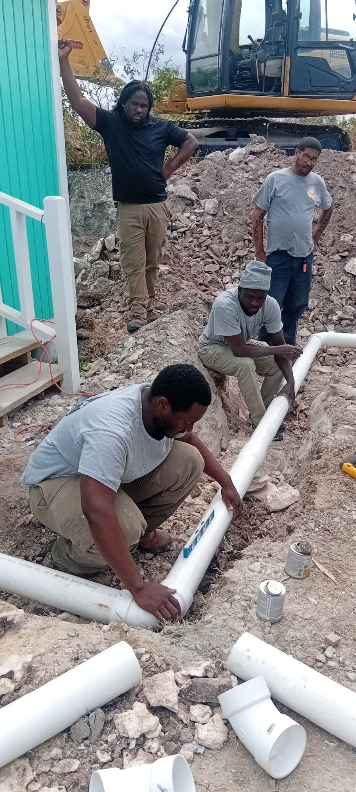 Five men working together to install a white PVC pipe in the ground near a teal-colored building. One man is crouching and holding the pipe, while another is sitting on the ground guiding it, and three others are standing nearby. There is constructio