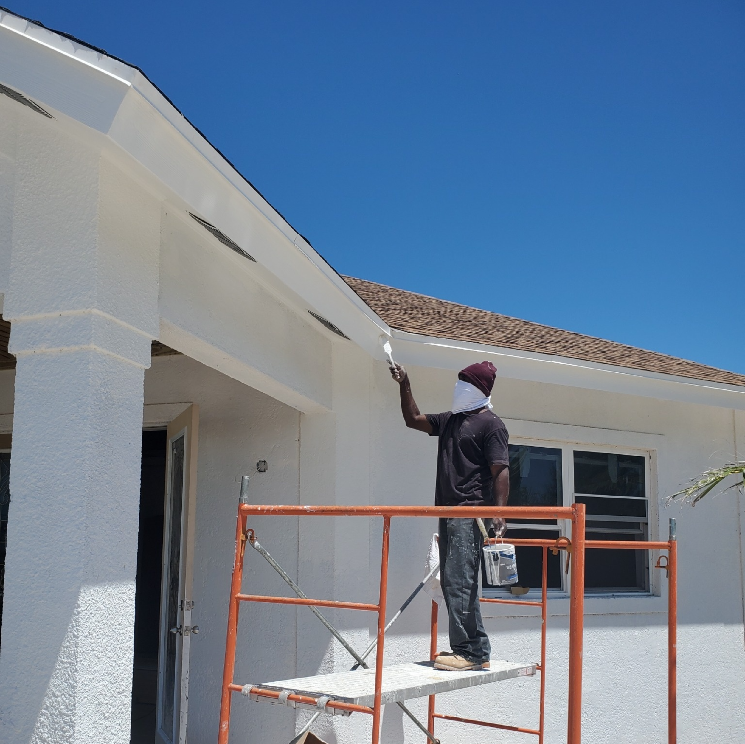 A person standing on scaffolding painting the exterior trim of a white house under a clear blue sky.
