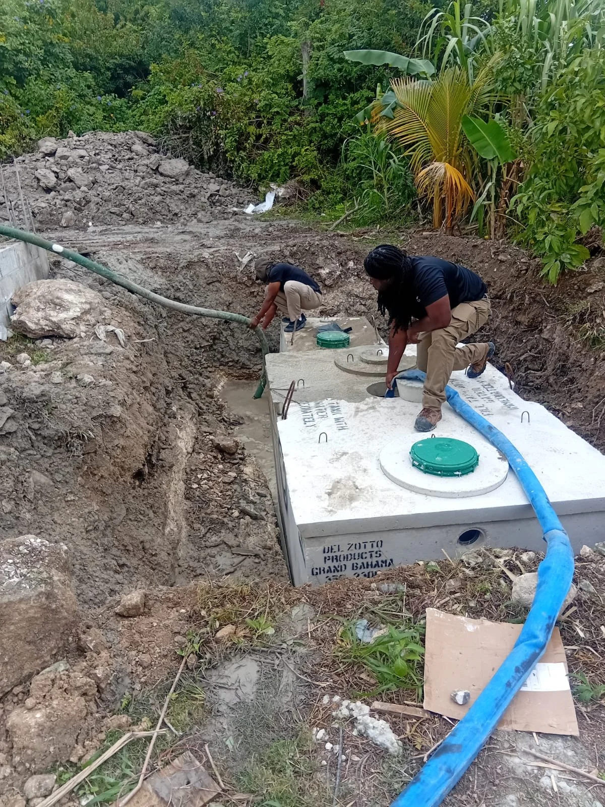 Two workers install a septic tank underground in a backyard with lush green foliage.