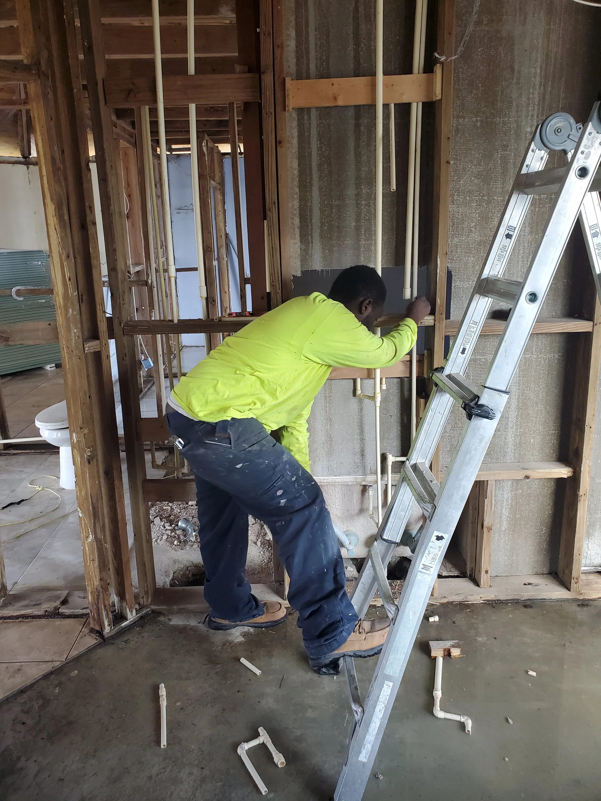 A worker in a yellow shirt installing plumbing pipes in a building under construction, standing on a ladder.