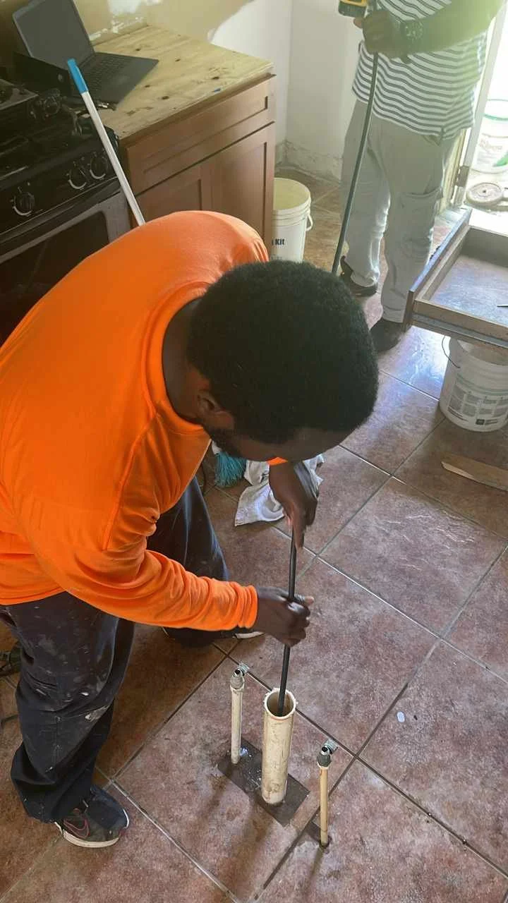 A man in an orange shirt is working on a plumbing pipe in a kitchen or utility room, with another person standing nearby. The man appears to be installing or repairing a pipe at the floor level.