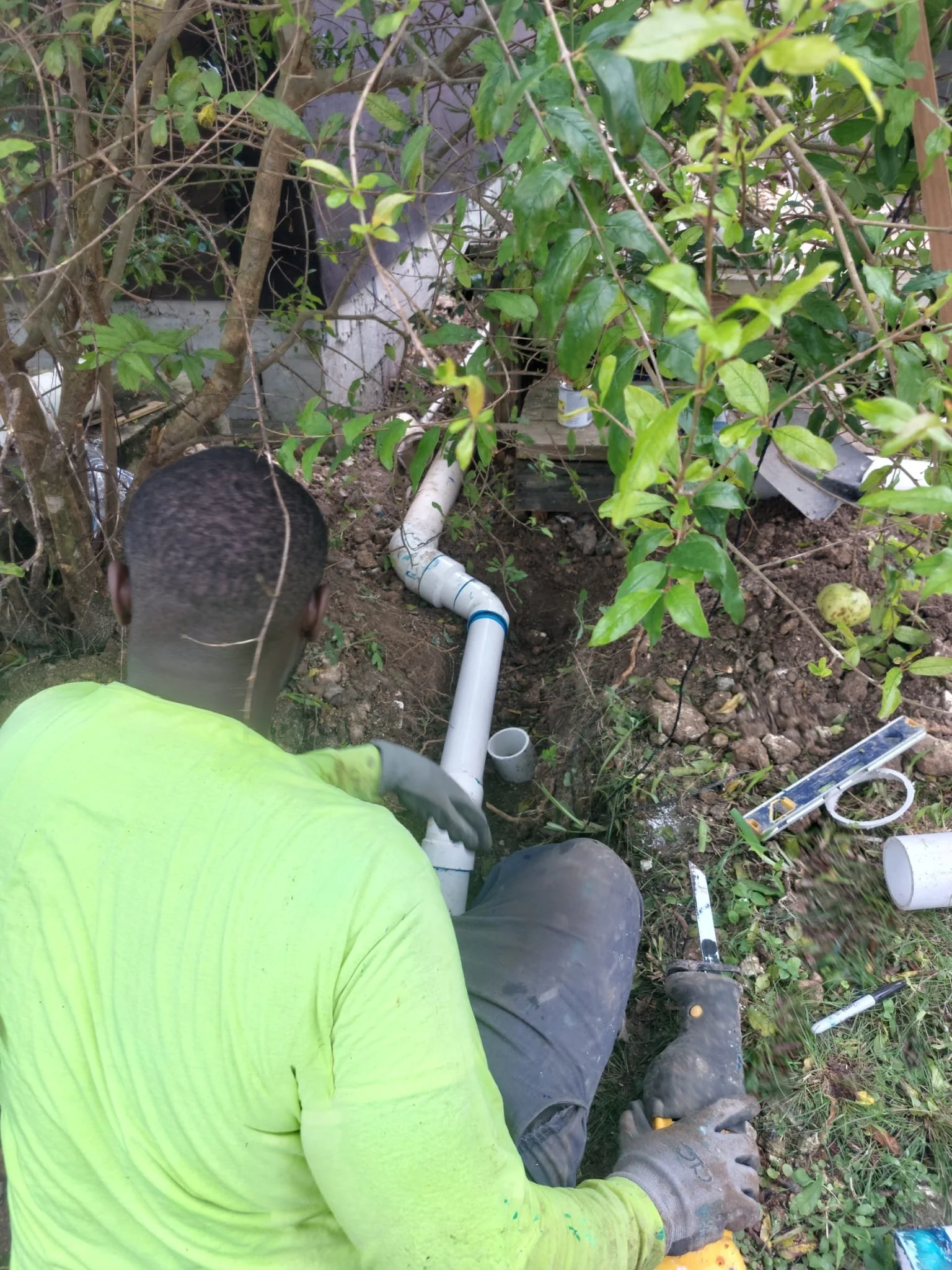 A person working on sewer pipe repairs outdoors among bushes, using tools like a pipe cutter and level, wearing gloves and a bright yellow shirt.