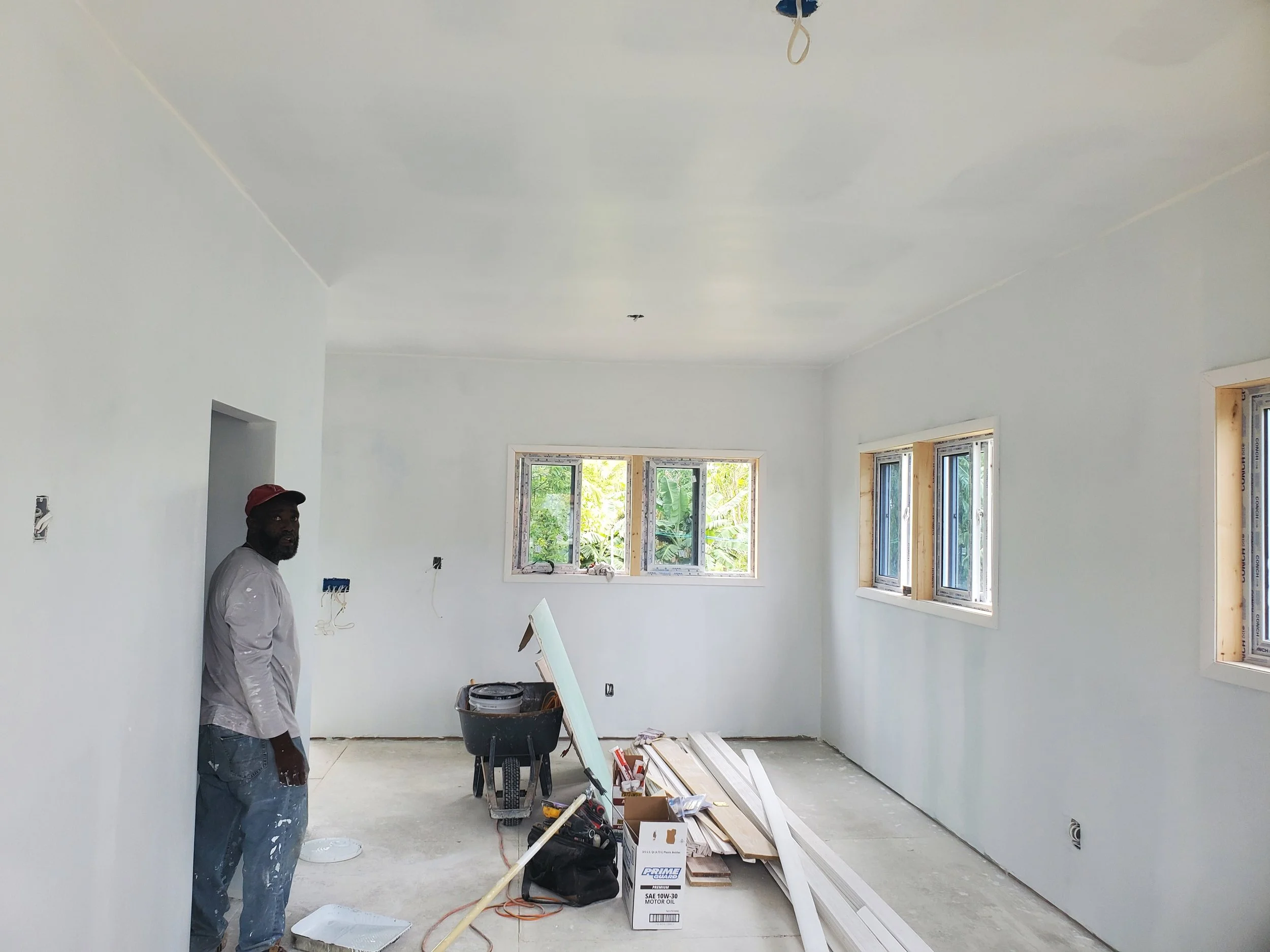 Interior of a room under construction with a worker standing near the wall, windows installed, and construction tools and materials on the floor.